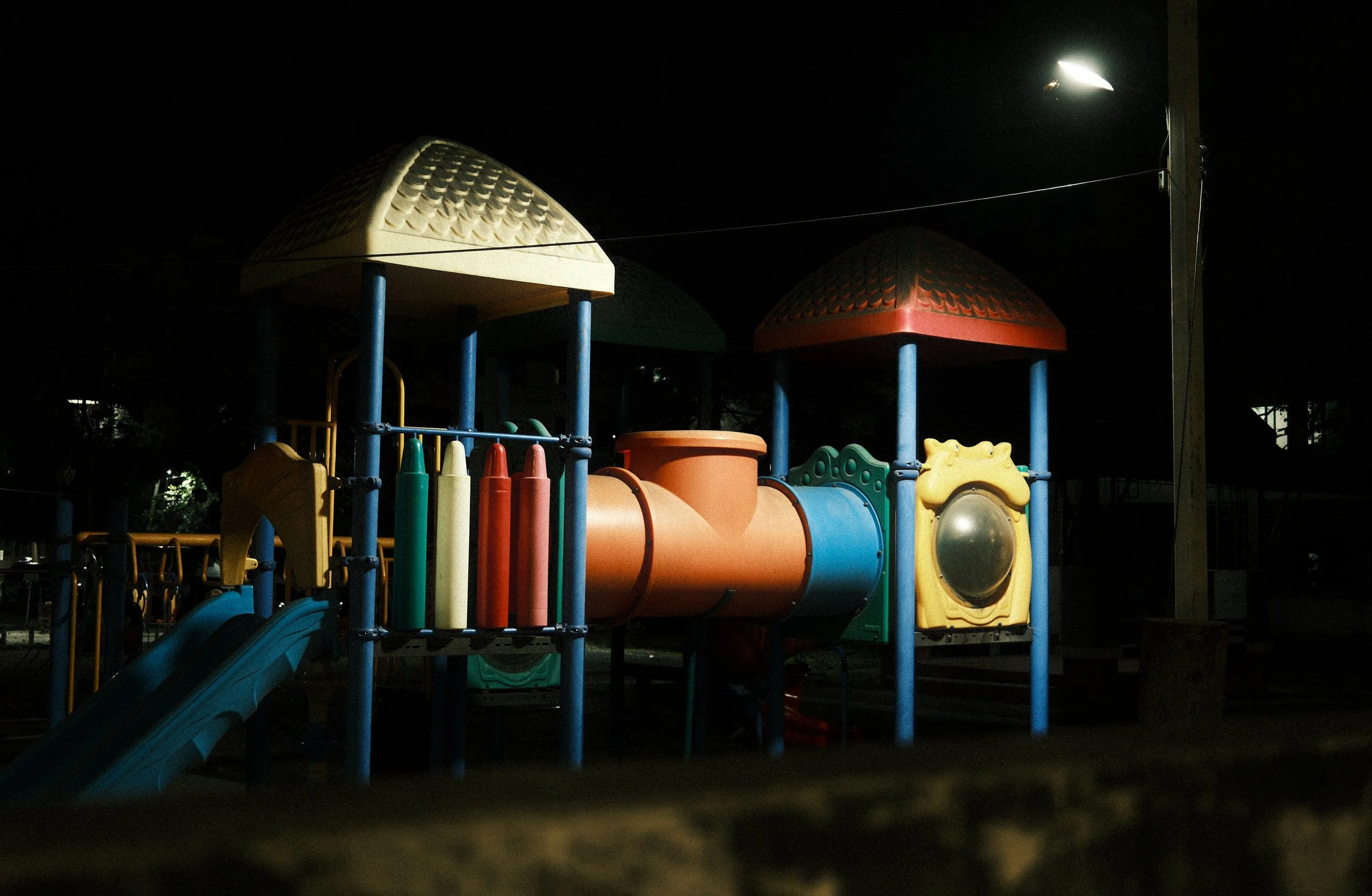 Colorful playground structure at night with slide, tubes, and towers, illuminated by a nearby street lamp.