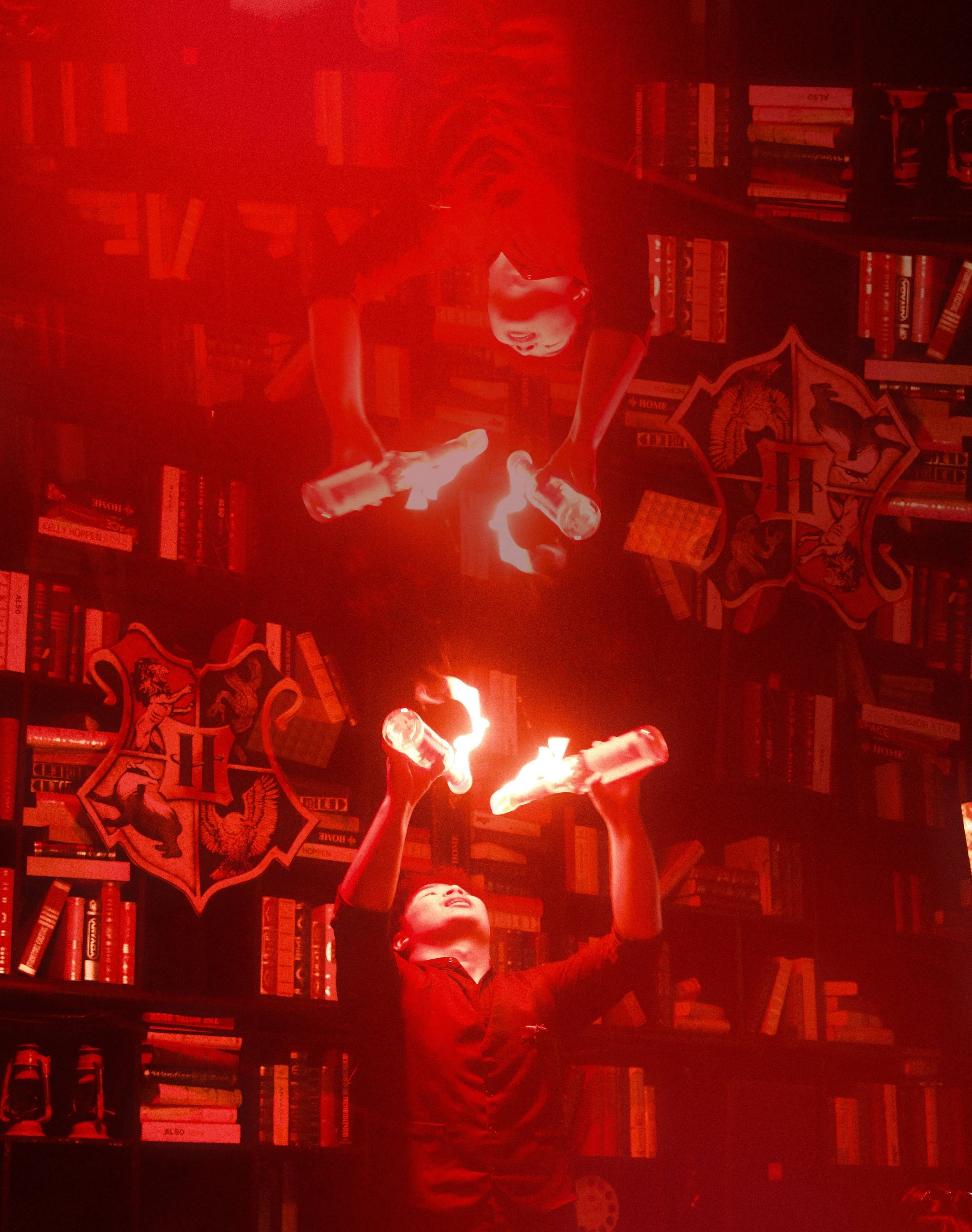 A person in a black shirt juggling three bottles illuminated by red light, with a bookshelf and a Harry Potter crest on the wall behind them.