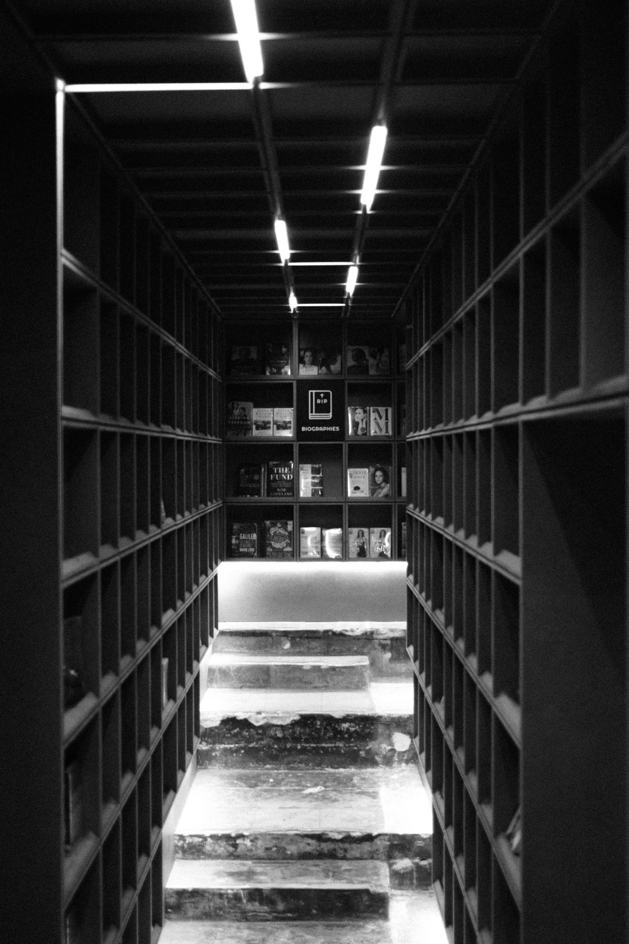 View down a narrow aisle with shelves of books on both sides, leading to a display wall at the end with additional books and magazine covers, illuminated by overhead strip lighting, in black and white.