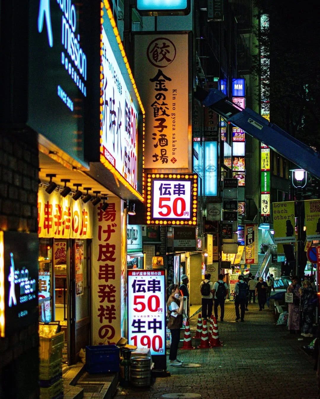 Nighttime street scene in Japan with neon signs in Japanese, street vendors, and pedestrians walking along the sidewalk.
