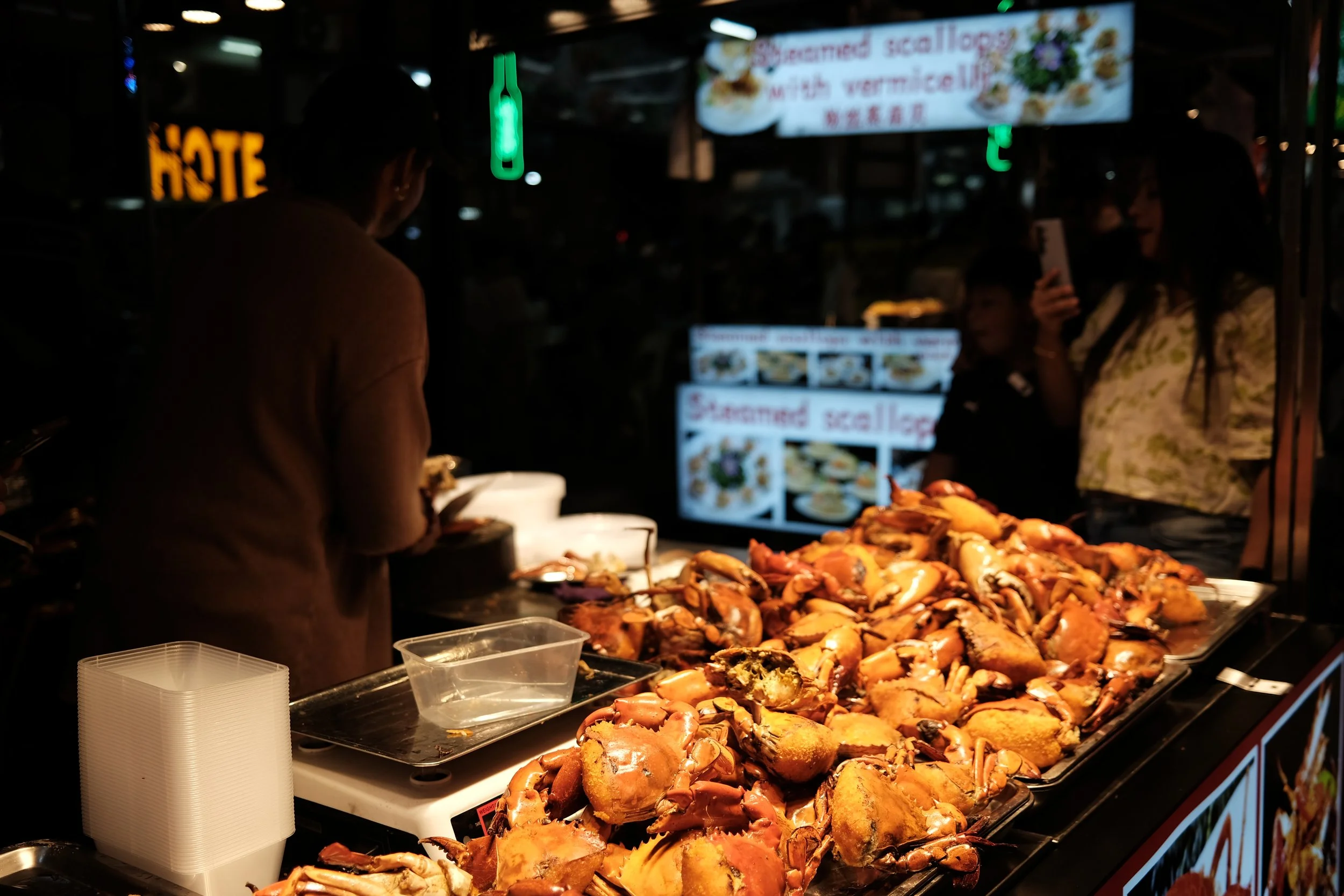 Fresh cooked crabs displayed on a food stall at night, with people in the background and illuminated signs.