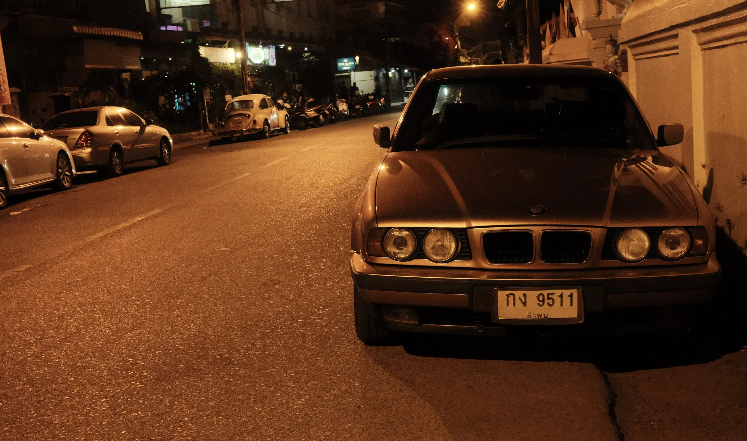 A street scene at night with several parked cars along the roadside, including an older model gray BMW in the foreground. The street is dimly lit with streetlights and illuminated signs in the background. The area appears to be in an urban setting wi