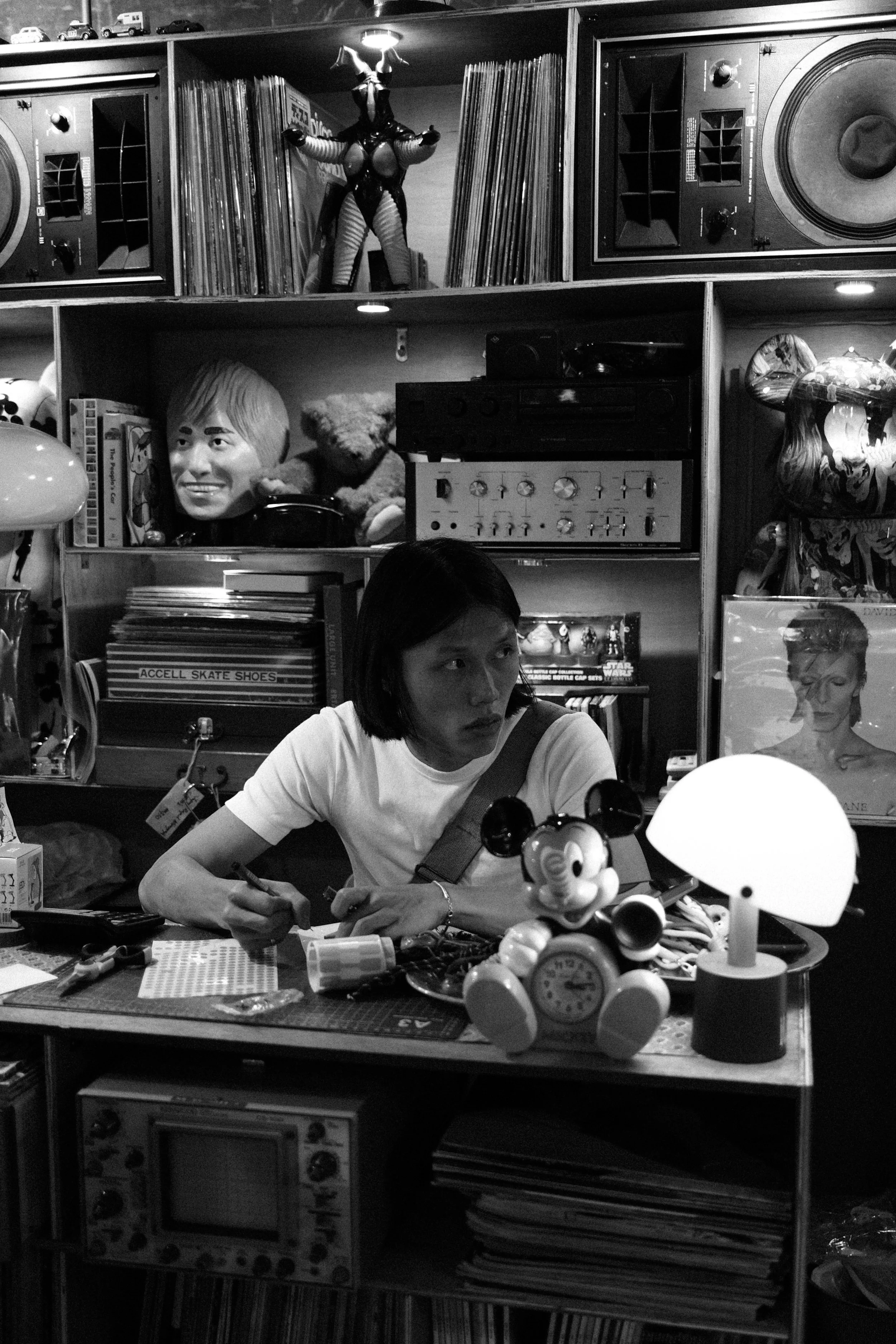 A woman sitting at a cluttered table in a room filled with various collectibles, toys, books, and art pieces. She is looking to her side with a serious expression. The background contains shelves with records, a vintage radio, and decorative items.