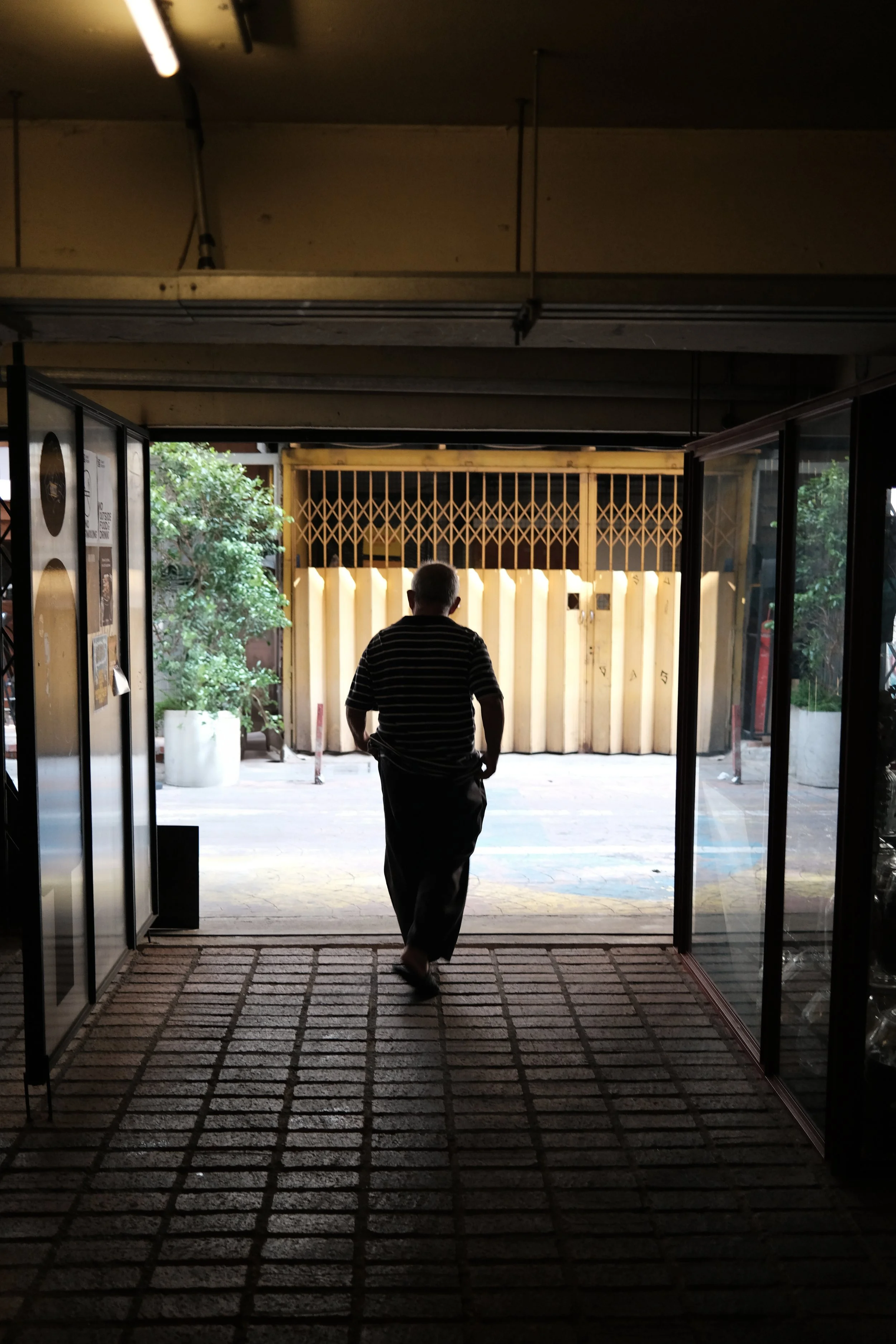 A person walking out of a building toward the street, silhouetted by sunlight outside, with trees and a closed gate visible.