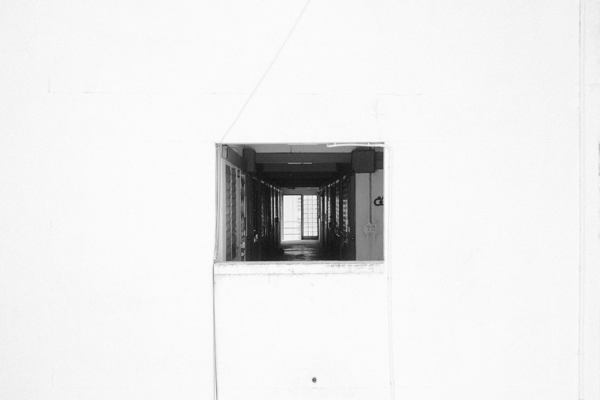Black and white photo of a corridor seen through a square opening in a white wall, with a door at the end of the corridor.