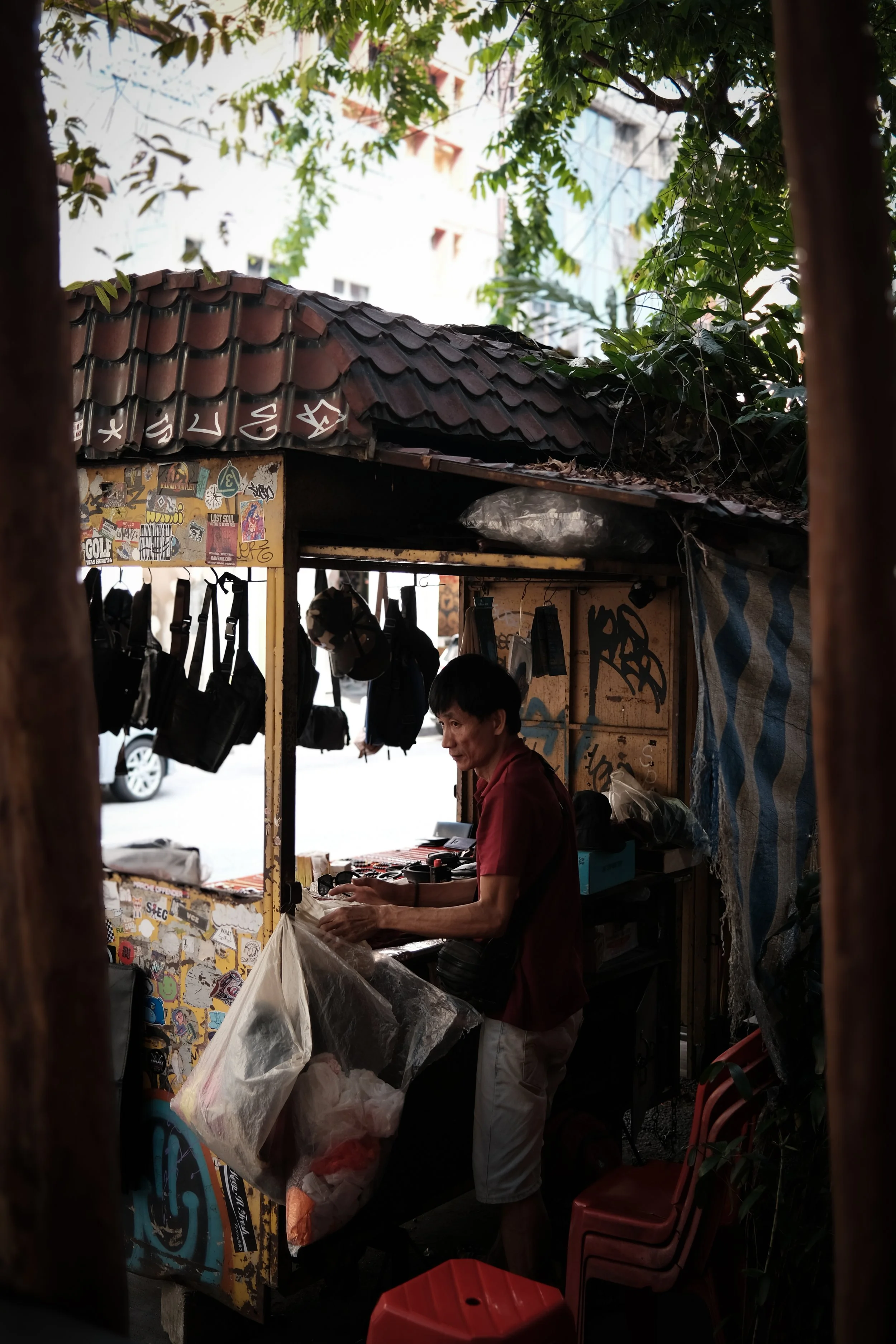 A man working at a small roadside shop with hanging bags and stickers on the stall, surrounded by trees and parked cars in the background.