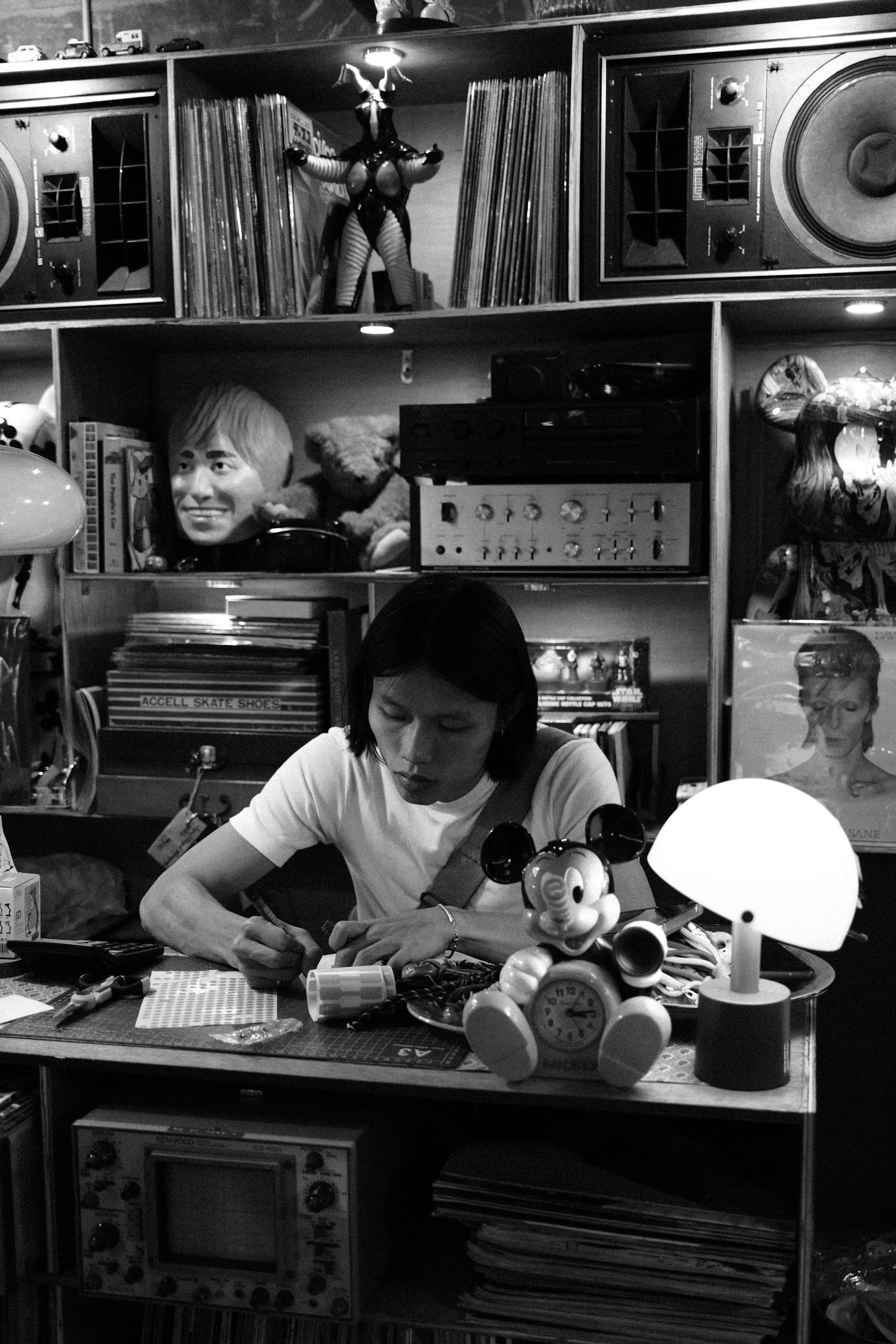 A woman sitting at a cluttered desk in a vintage store or room, surrounded by toys, records, and artwork, with shelves filled with books, vinyl records, and eclectic decorations, including a Mickey Mouse clock.