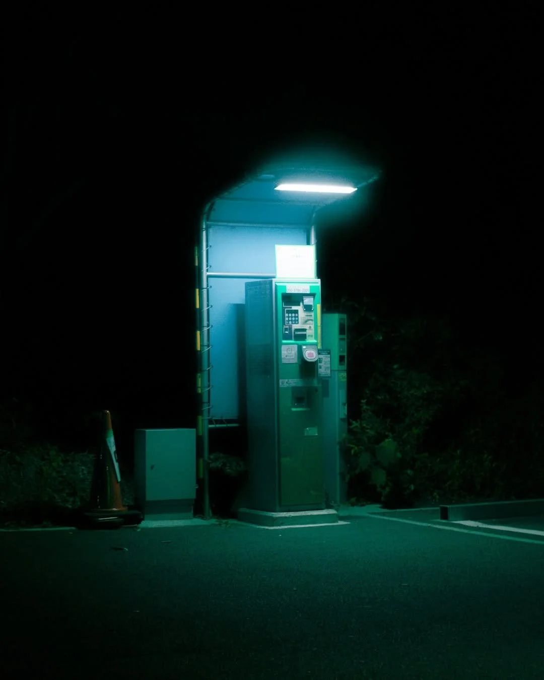 A parking lot booth illuminated by a blue-green fluorescent light at night, with traffic cones placed nearby.