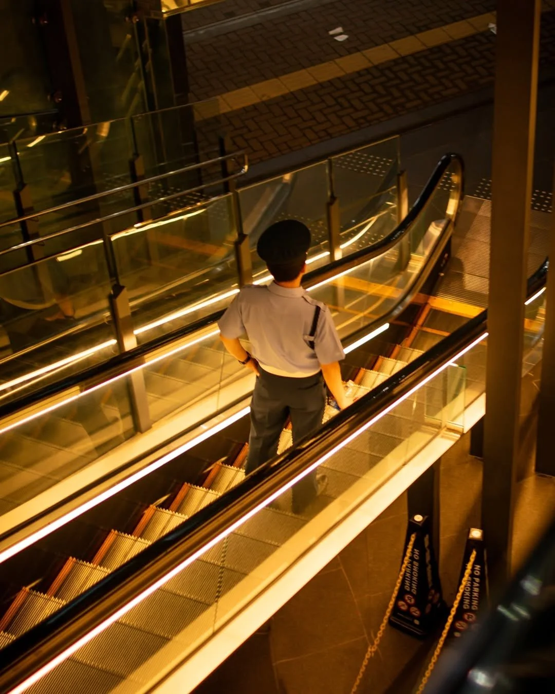 A security guard in uniform standing on an escalator in a well-lit indoor setting, with reflective glass panels and a 'No Dabbing' warning sign on the floor.