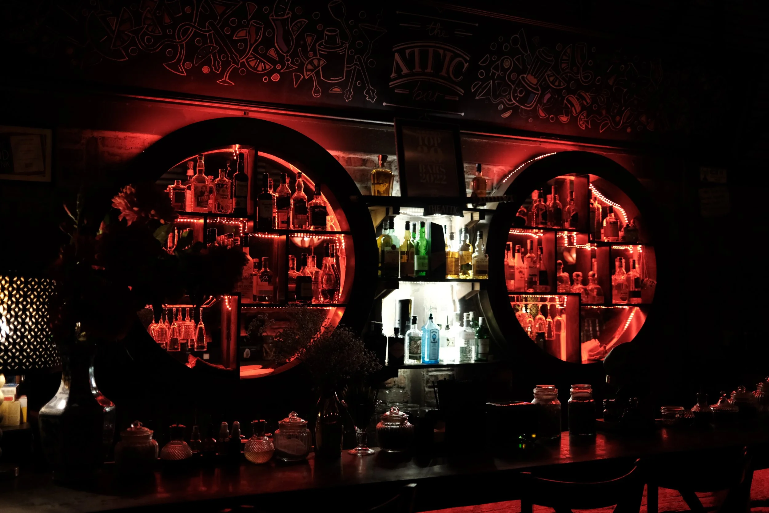 Dimly lit bar with illuminated shelves holding various bottles of alcohol behind a counter.