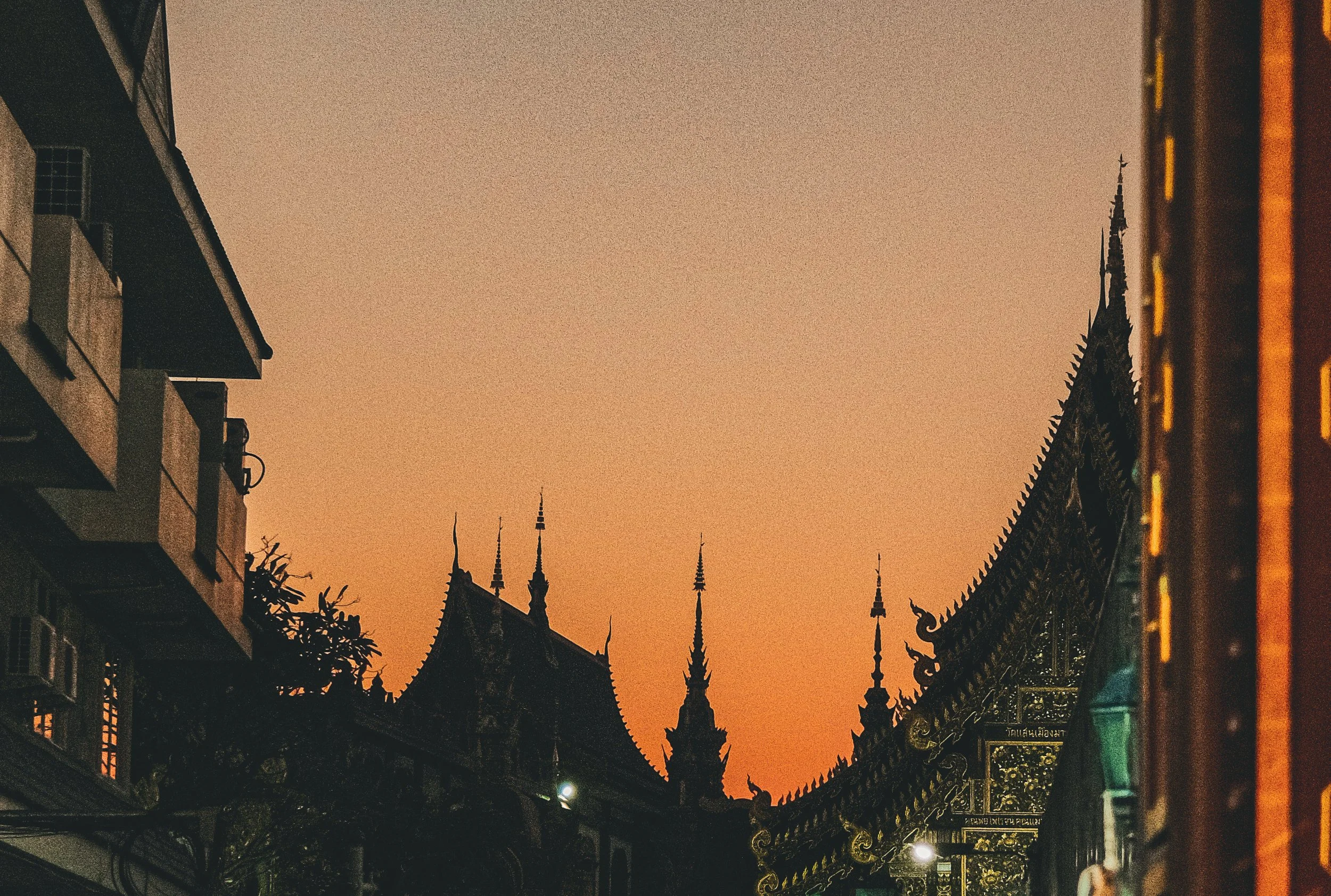 Silhouette of traditional Thai temple rooftops and spires at sunset, with nearby modern buildings.