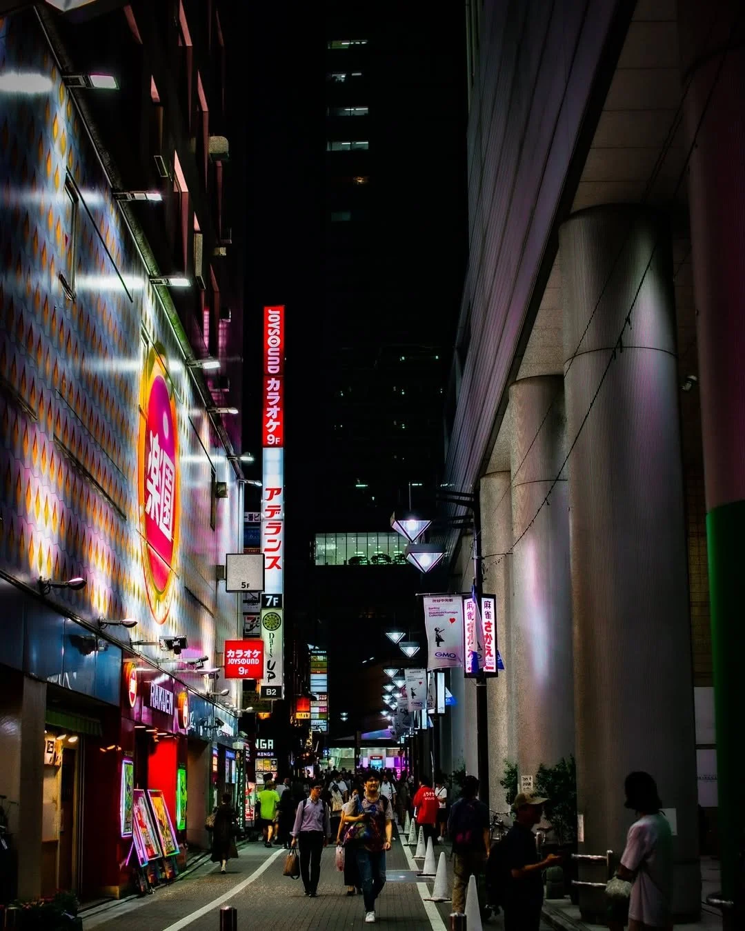 A vibrant city street at night with brightly lit signs in Japanese, pedestrians walking, and colorful storefronts, showcasing an urban nightlife scene.