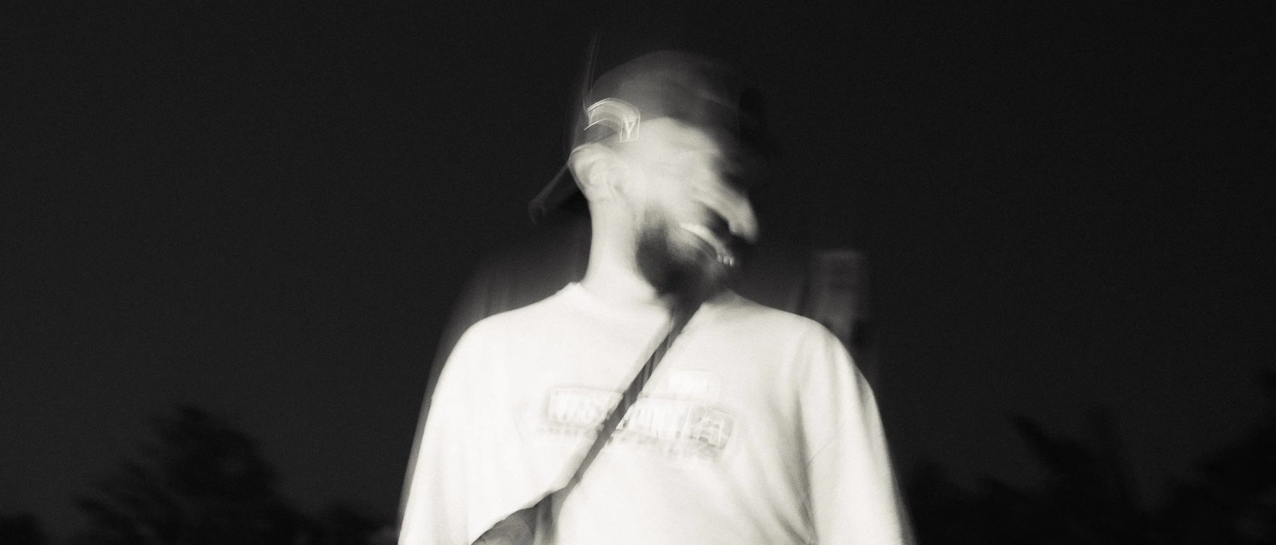 Black and white photo of a smiling man in a baseball cap and t-shirt, with motion blur creating a ghostly effect.