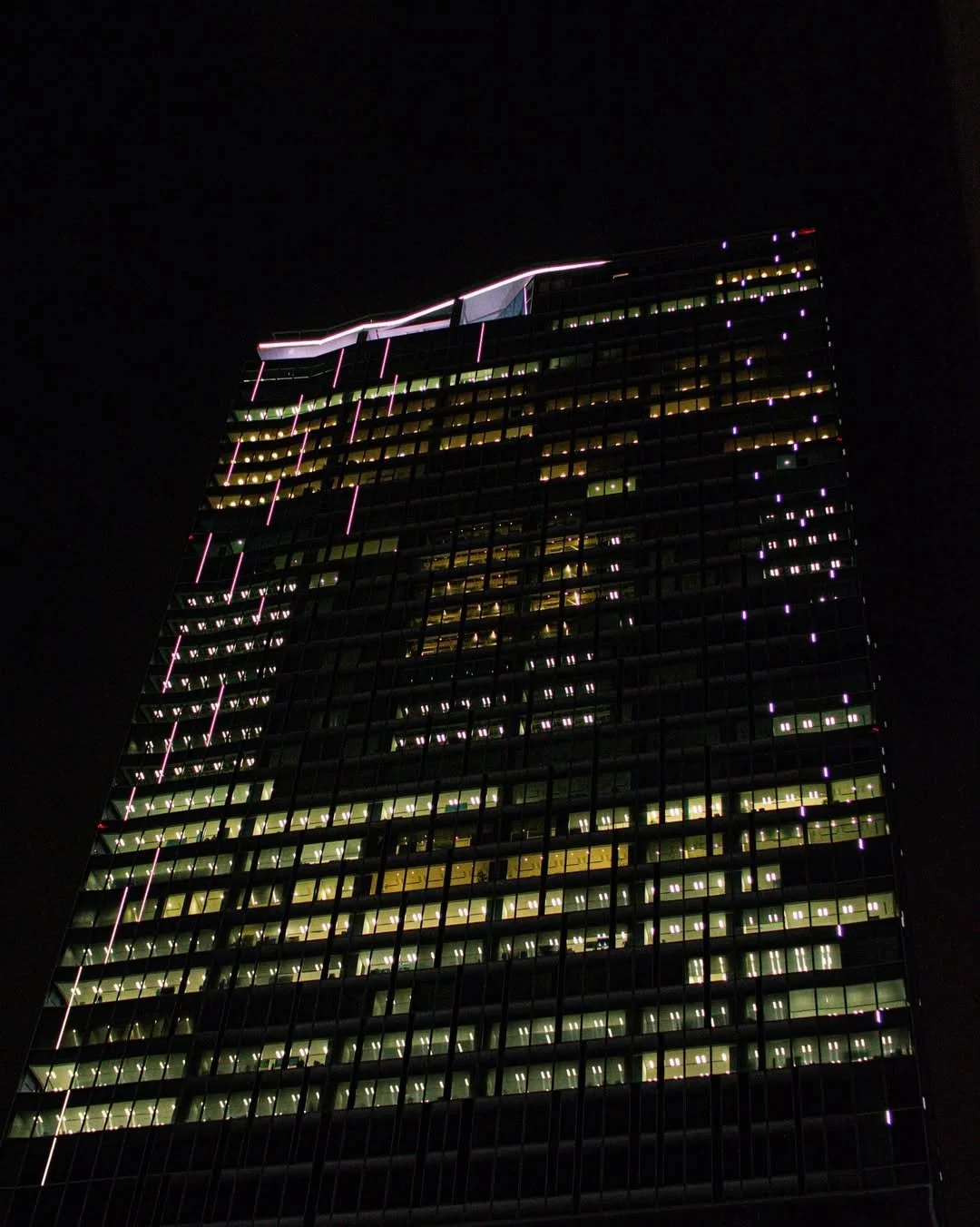 Night view of a tall office building illuminated with bright lights, with a dark sky behind it.