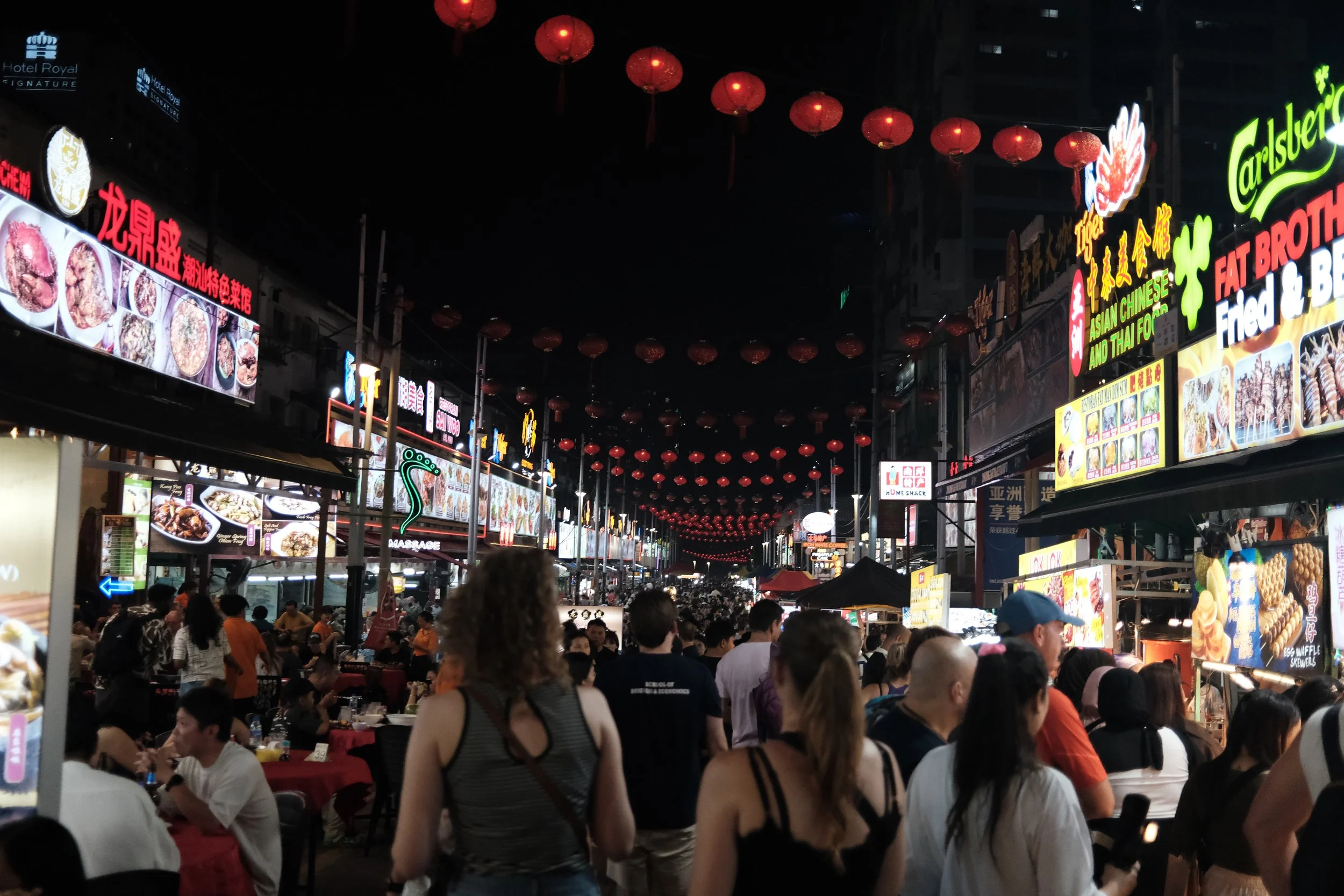 Night market street with illuminated signs in Chinese and English, crowded with people, and red lanterns hanging overhead.