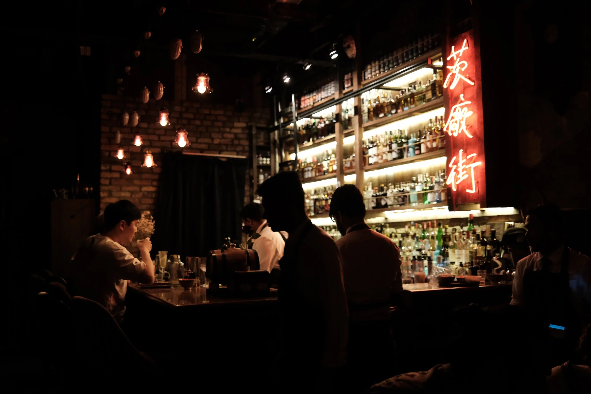 A dimly lit bar with a brick wall, illuminated bottles on shelves, and four people sitting and standing around the bar counter.