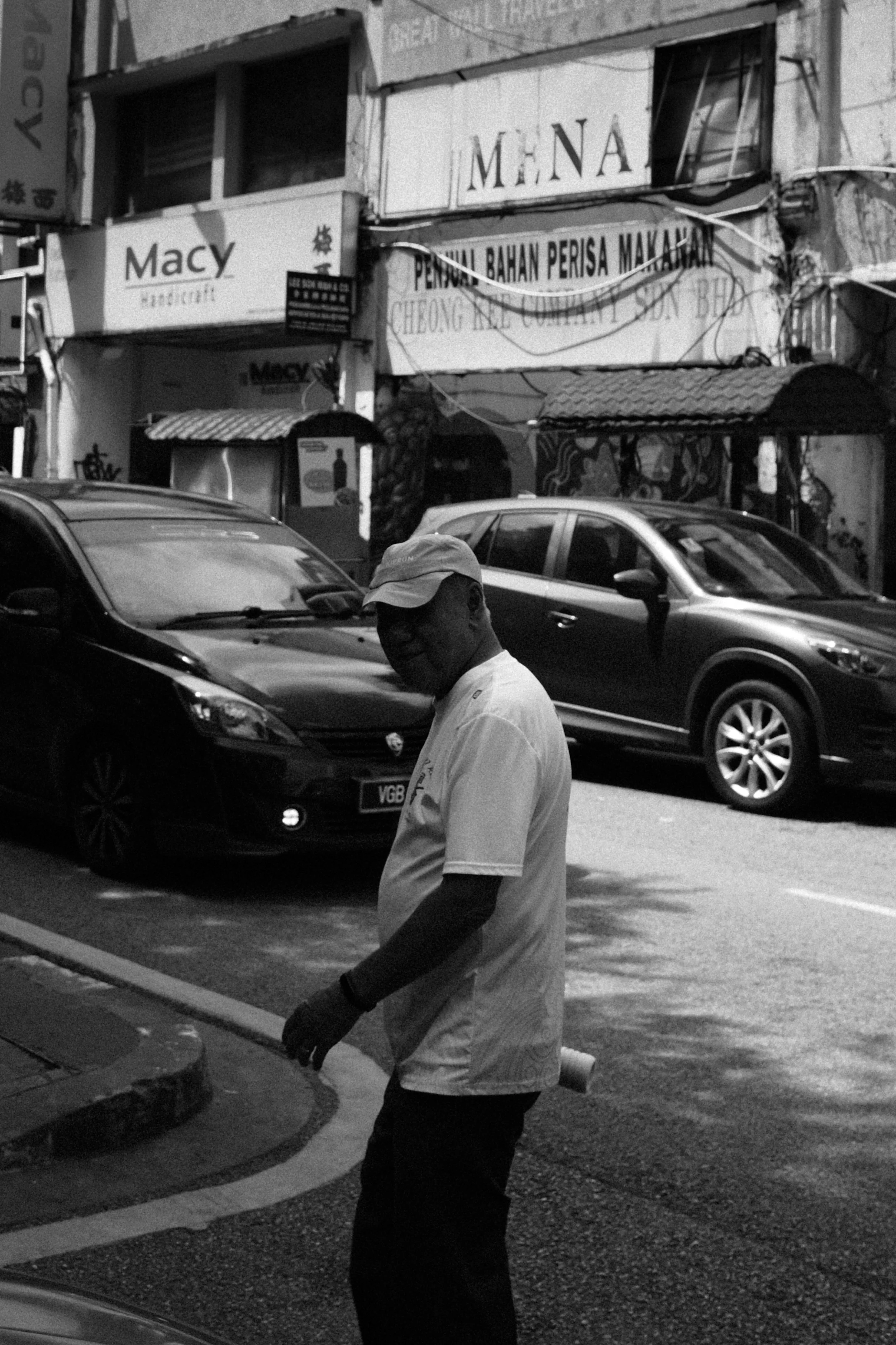 A man wearing a cap and white shirt standing on a street with cars parked behind him and storefronts with signs in a foreign language in the background.