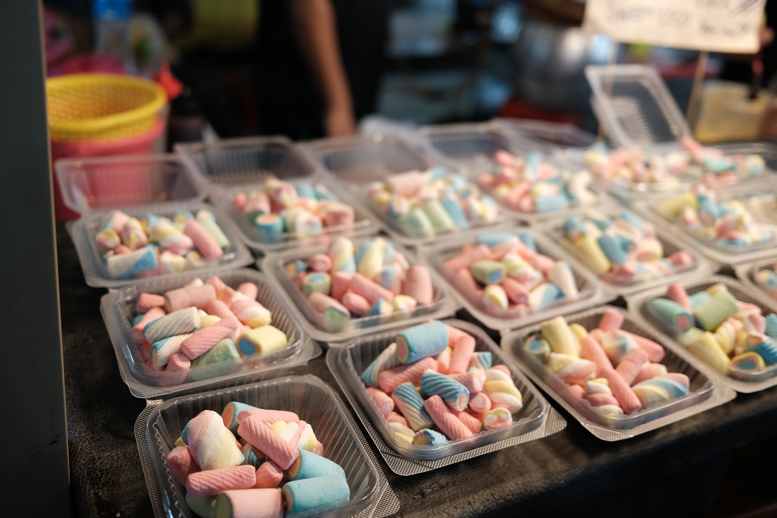Multiple clear plastic containers filled with colorful marshmallows on a dark table at a market stall.