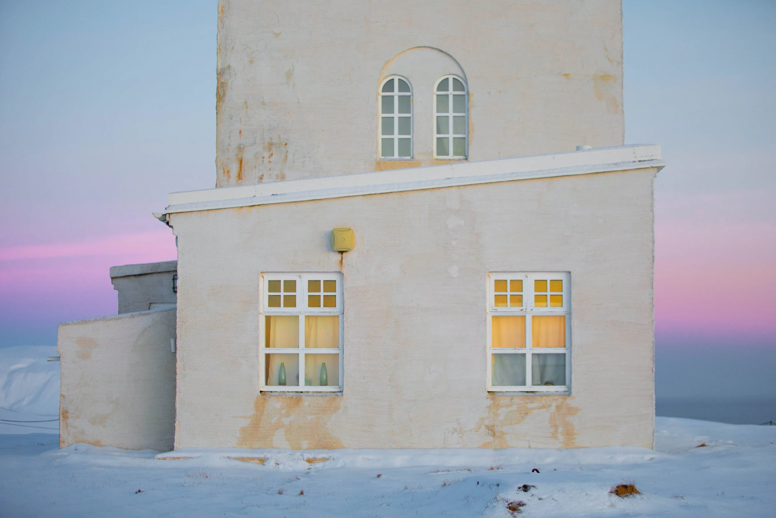 White Arctic building with glowing windows surrounded by snow beneath a pastel sunset sky