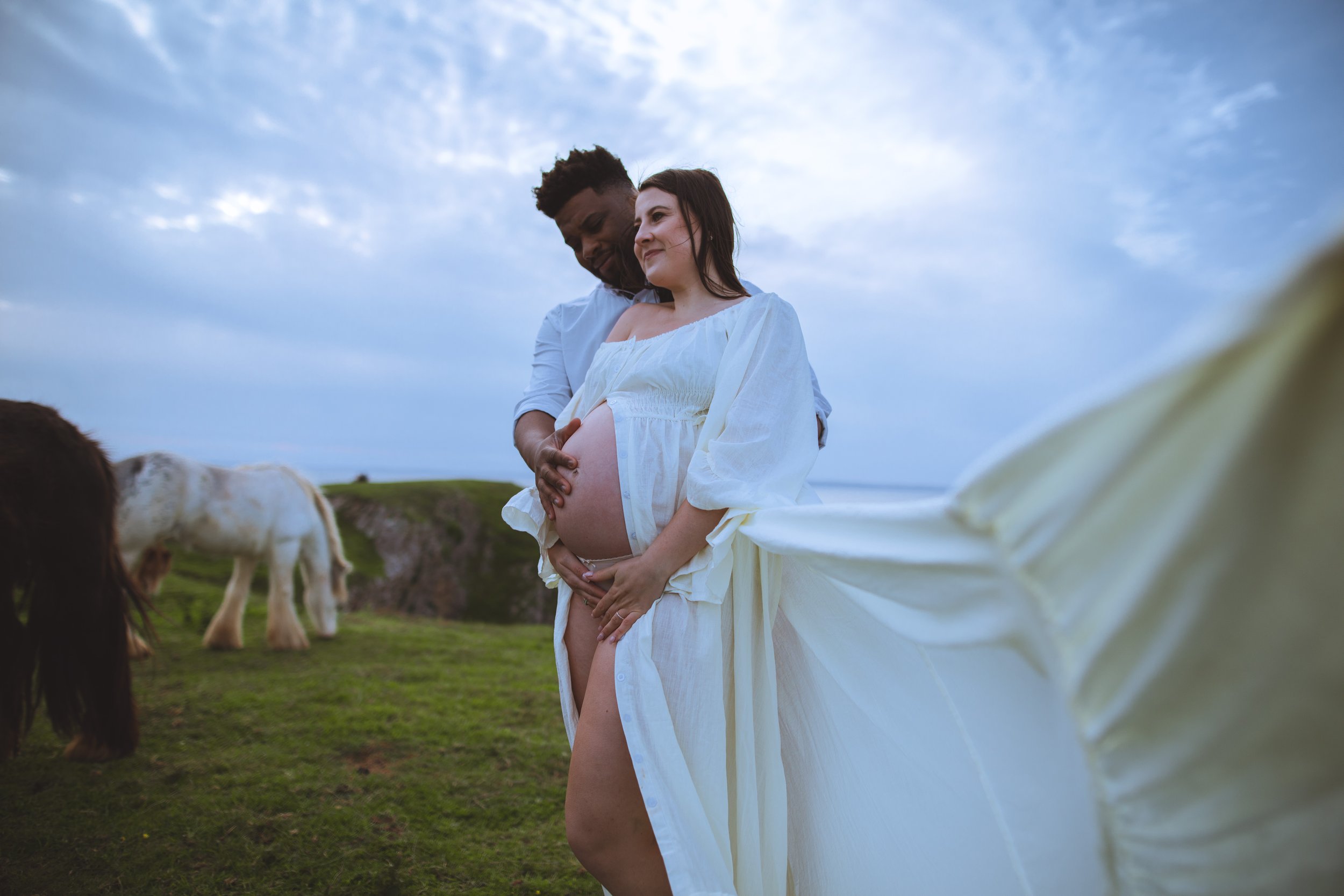 A pregnant woman in a white dress standing outdoors on a grassy hillside under a cloudy sky, holding her belly, with a man behind her touching her belly, and horses grazing in the background.