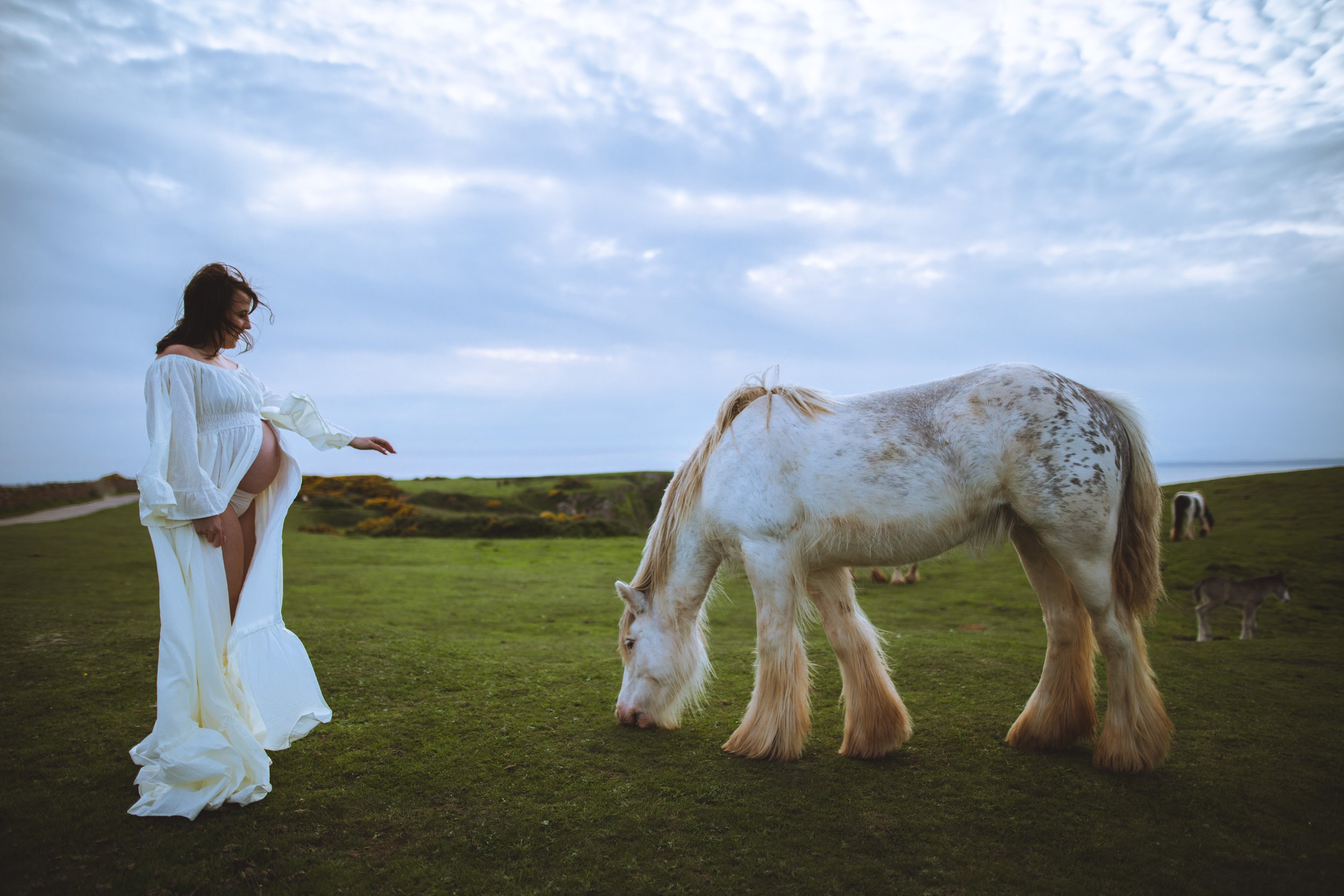 Pregnant woman in a long white dress standing on a grassy field, gently touching a white horse with brown spots grazing nearby, with a few other horses in the background and a cloudy sky overhead.