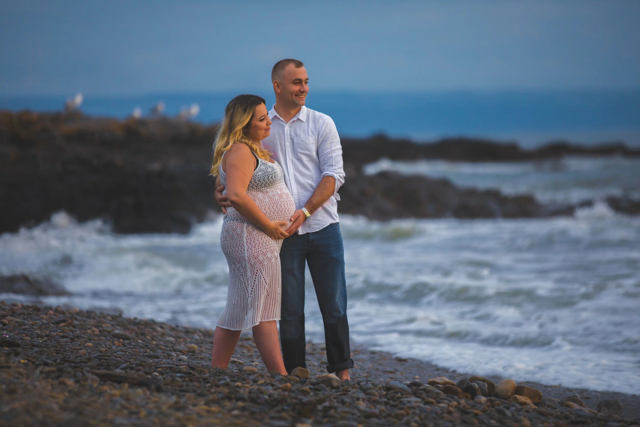 A couple standing on a rocky beach with ocean waves and a distant pier in the background, embracing and smiling, during dusk or early evening.