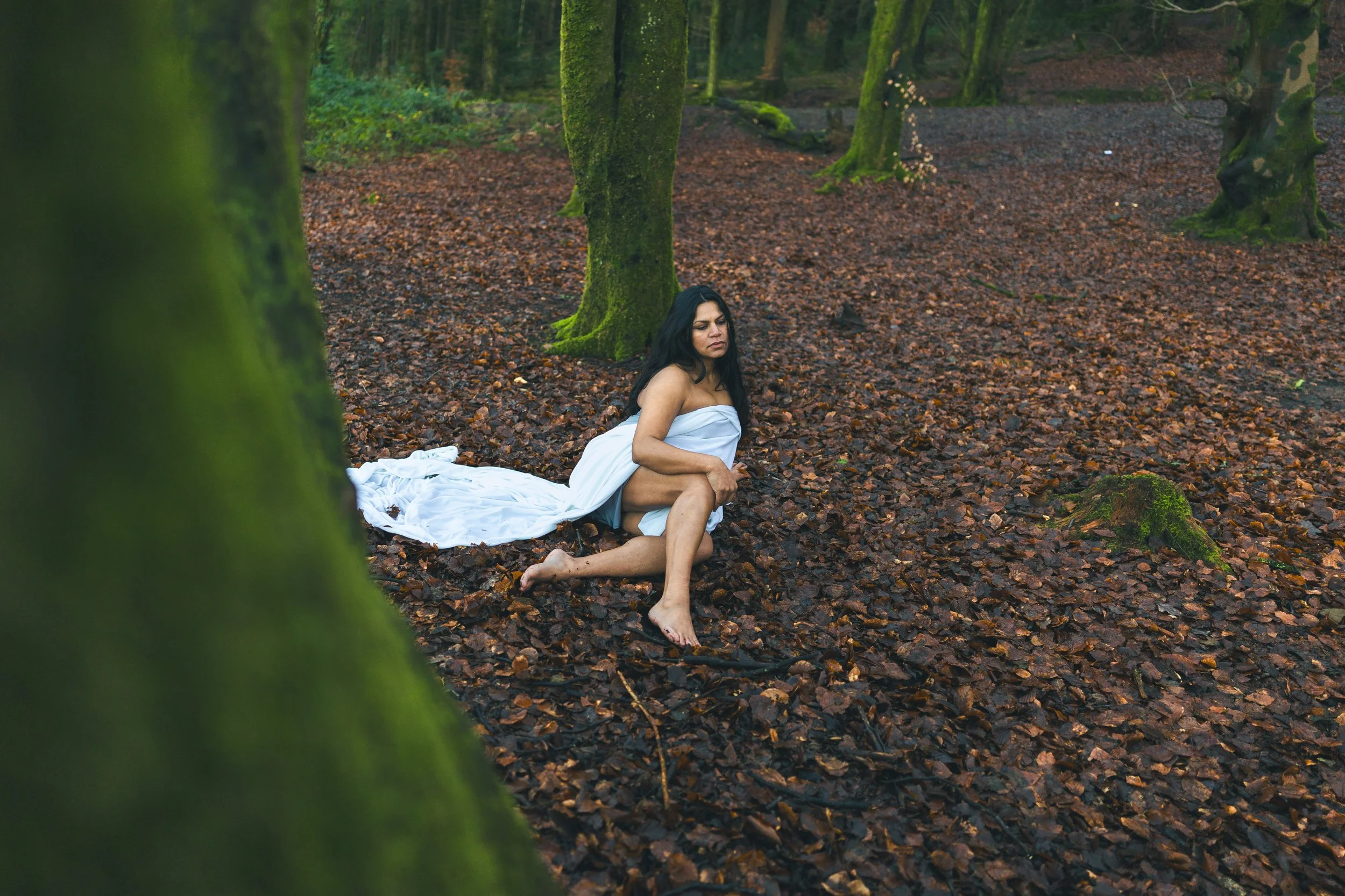 A woman with dark hair sits on the leaf-covered forest floor, wrapped in a white cloth, with a serious expression on her face. The scene is set in a dense, green woodland area.