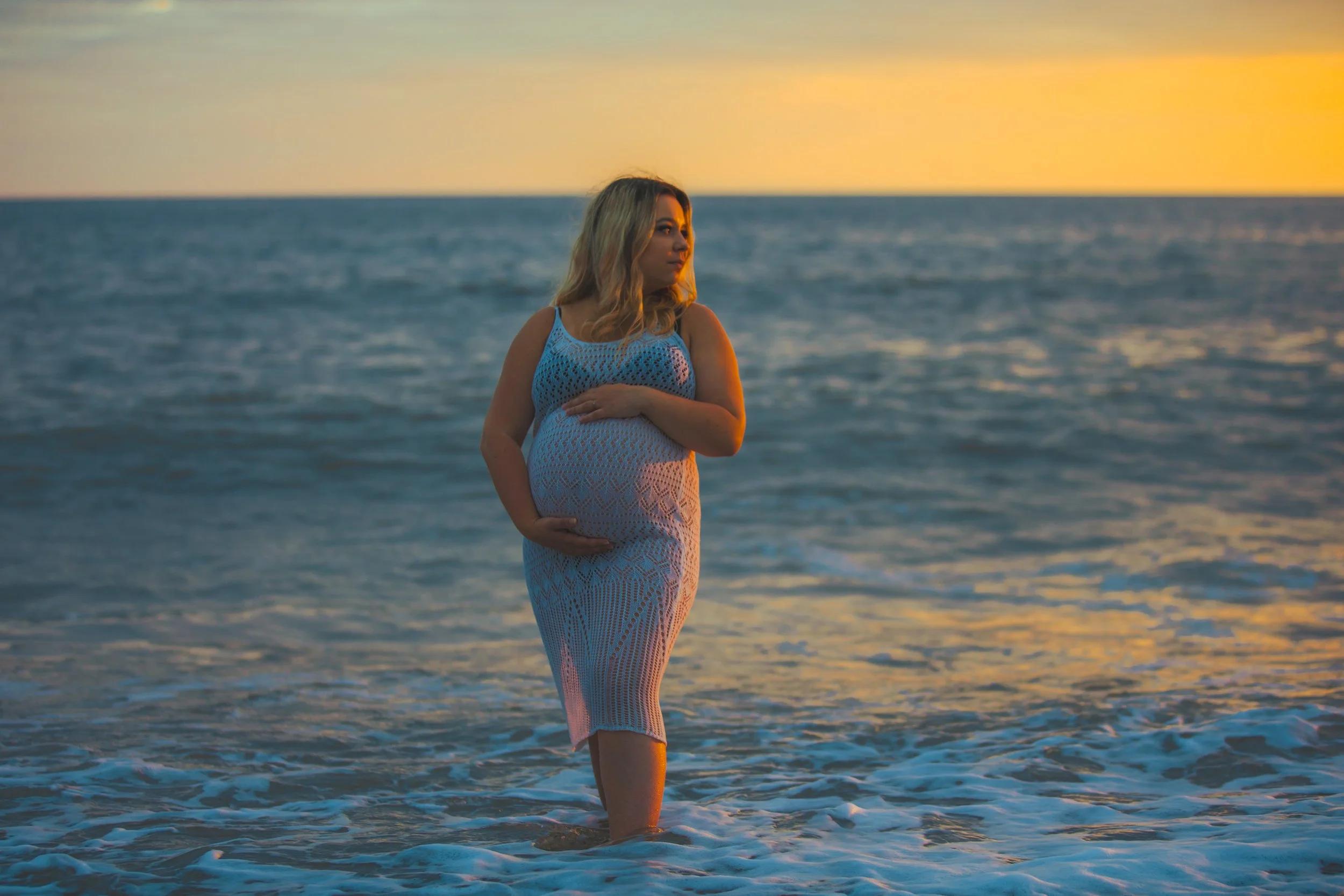 Pregnant woman in a white crochet dress standing in the ocean at sunset.