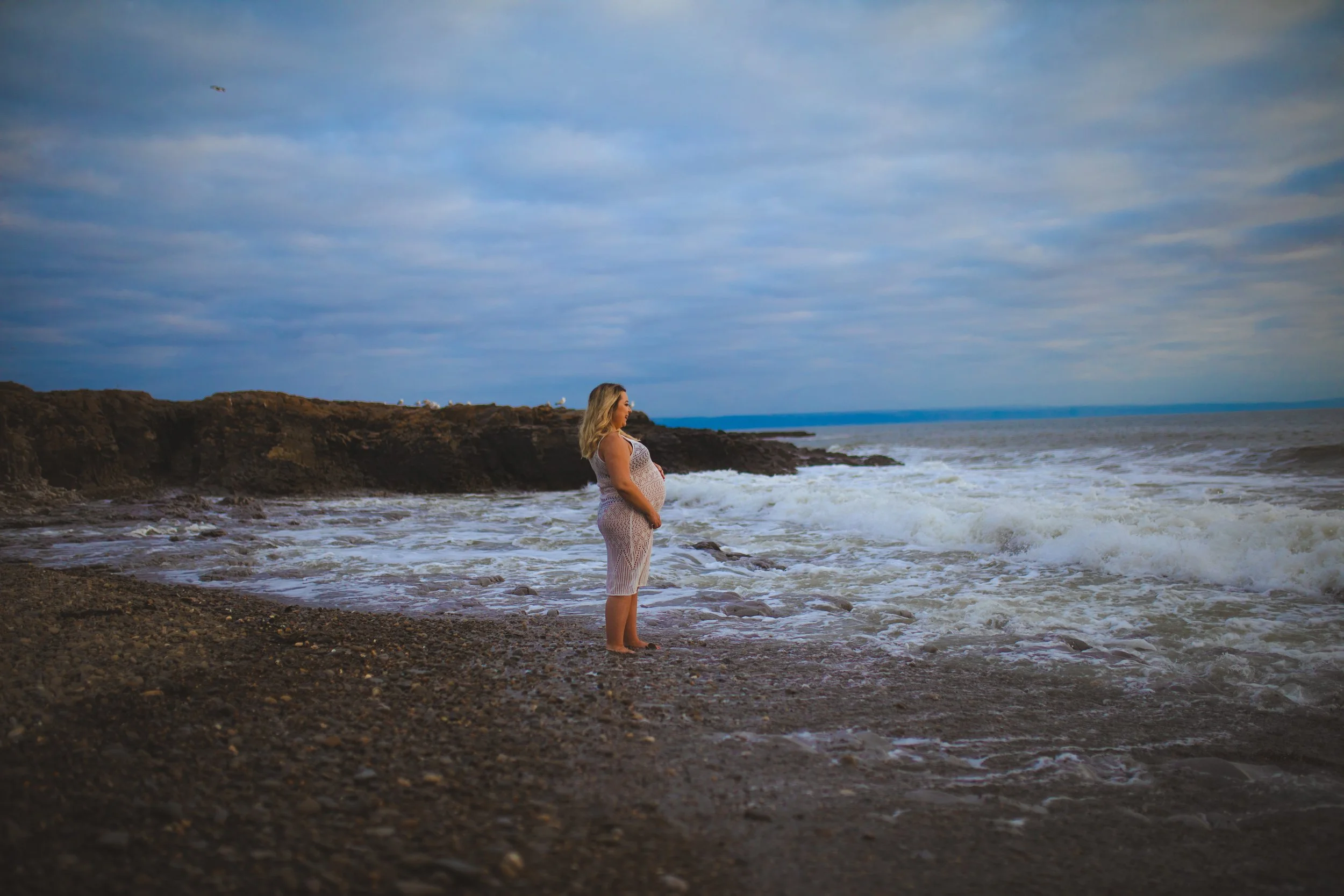 A pregnant woman standing barefoot on a rocky beach, gazing at the ocean waves under a cloudy sky.