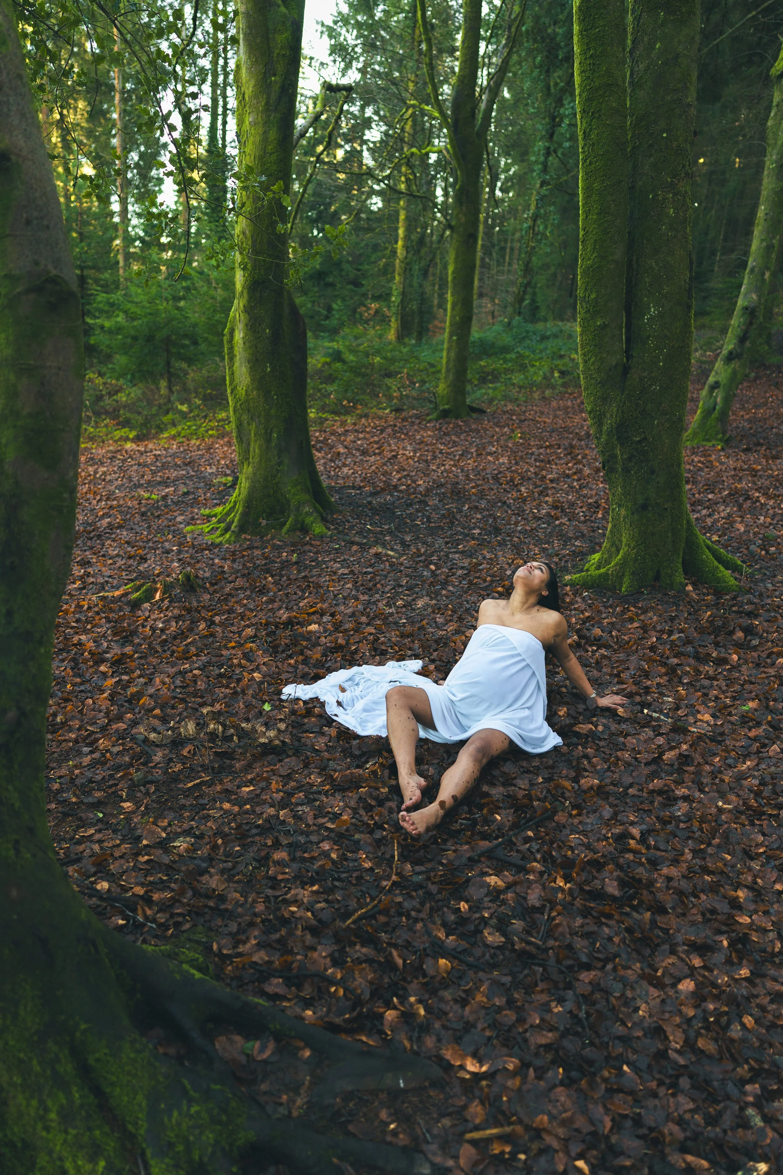 A woman in a white dress sitting on the forest floor among tall moss-covered trees, with her head tilted back and eyes closed, in a peaceful outdoor scene.