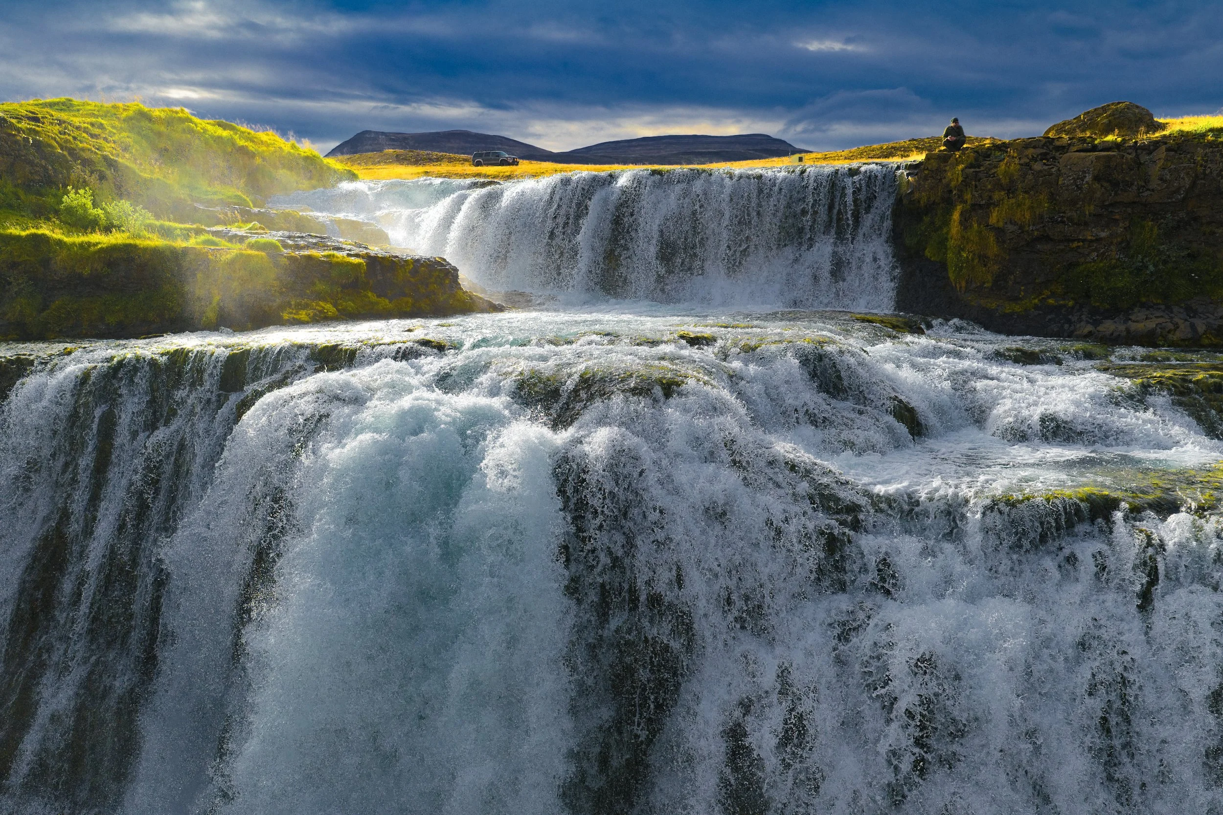 Panoramic Icelandic waterfall cascading over moss-covered basalt ledges beneath a cloudy sky, with mountains, a vehicle, and a person in the background.