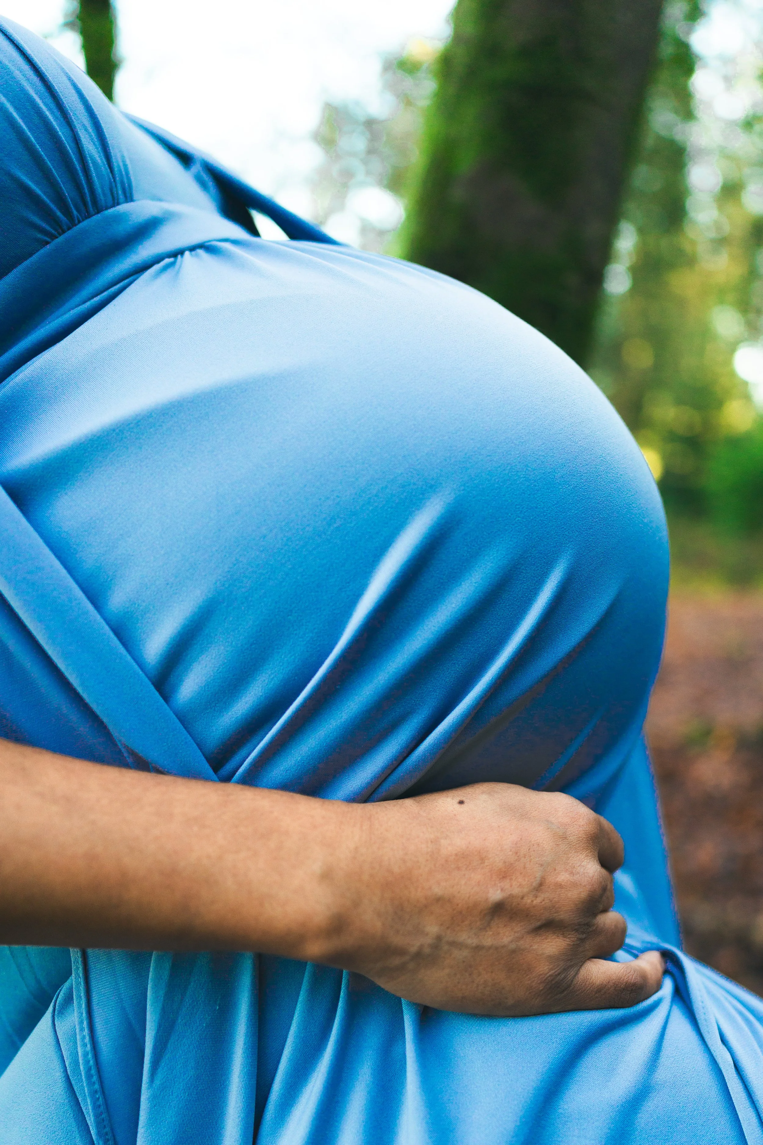 Close-up of a person holding their stomach while wearing a light blue shirt outdoors in a wooded area.