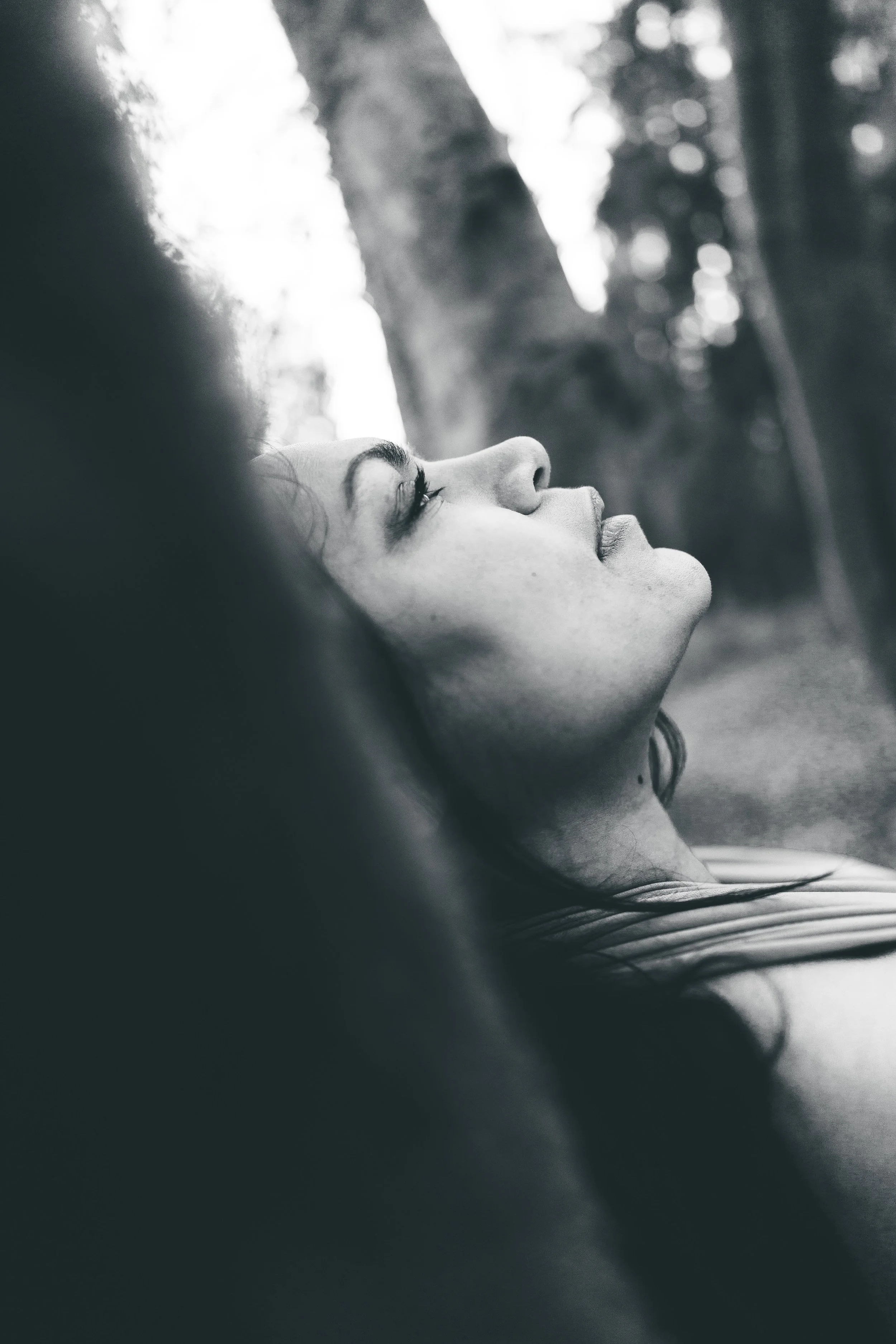A black and white photo of a woman lying outdoors with her eyes closed, facing upward, with trees in the background.