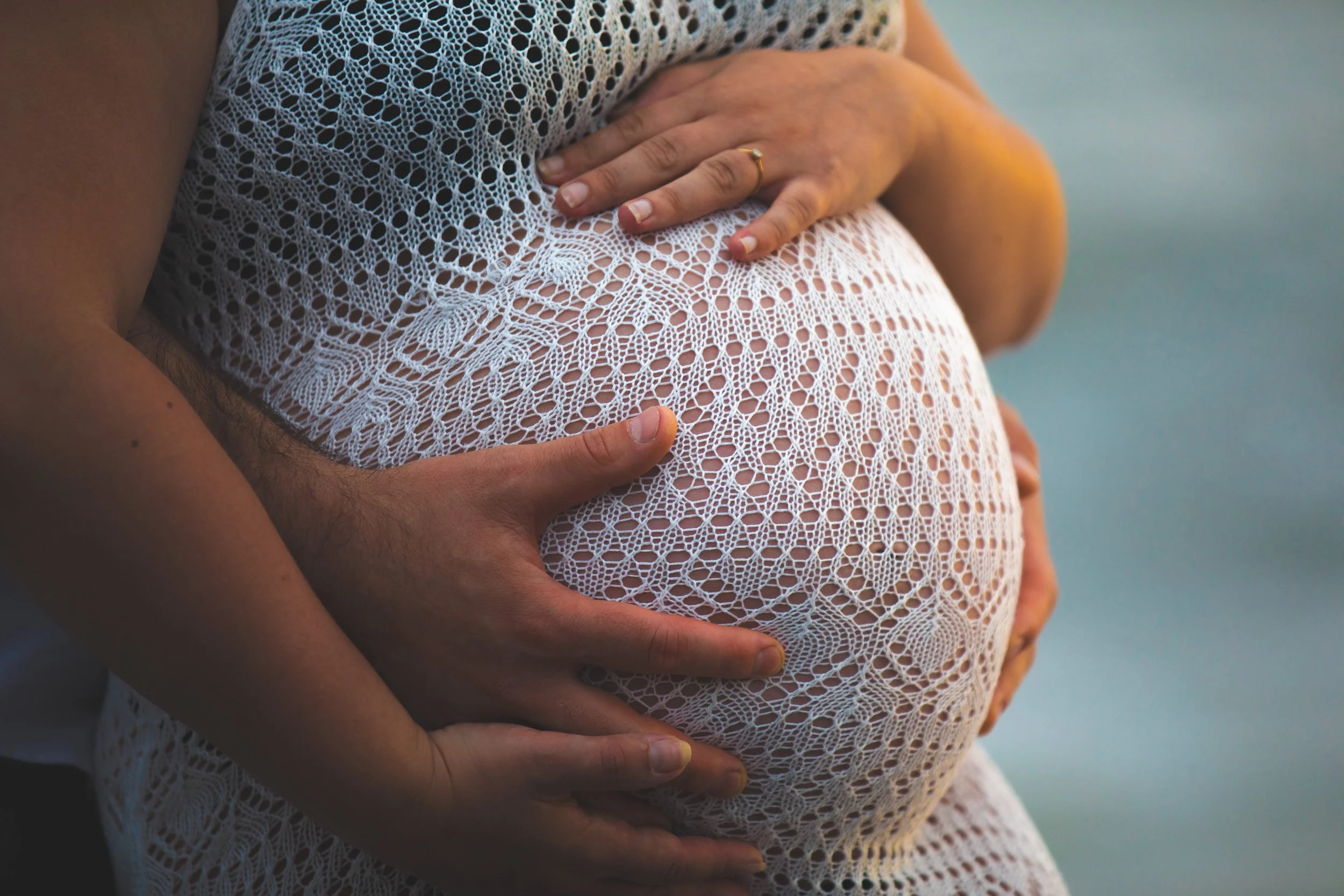 Close-up of a pregnant woman with a man holding her belly, wearing a white crochet dress with intricate patterns, by the water at sunset.