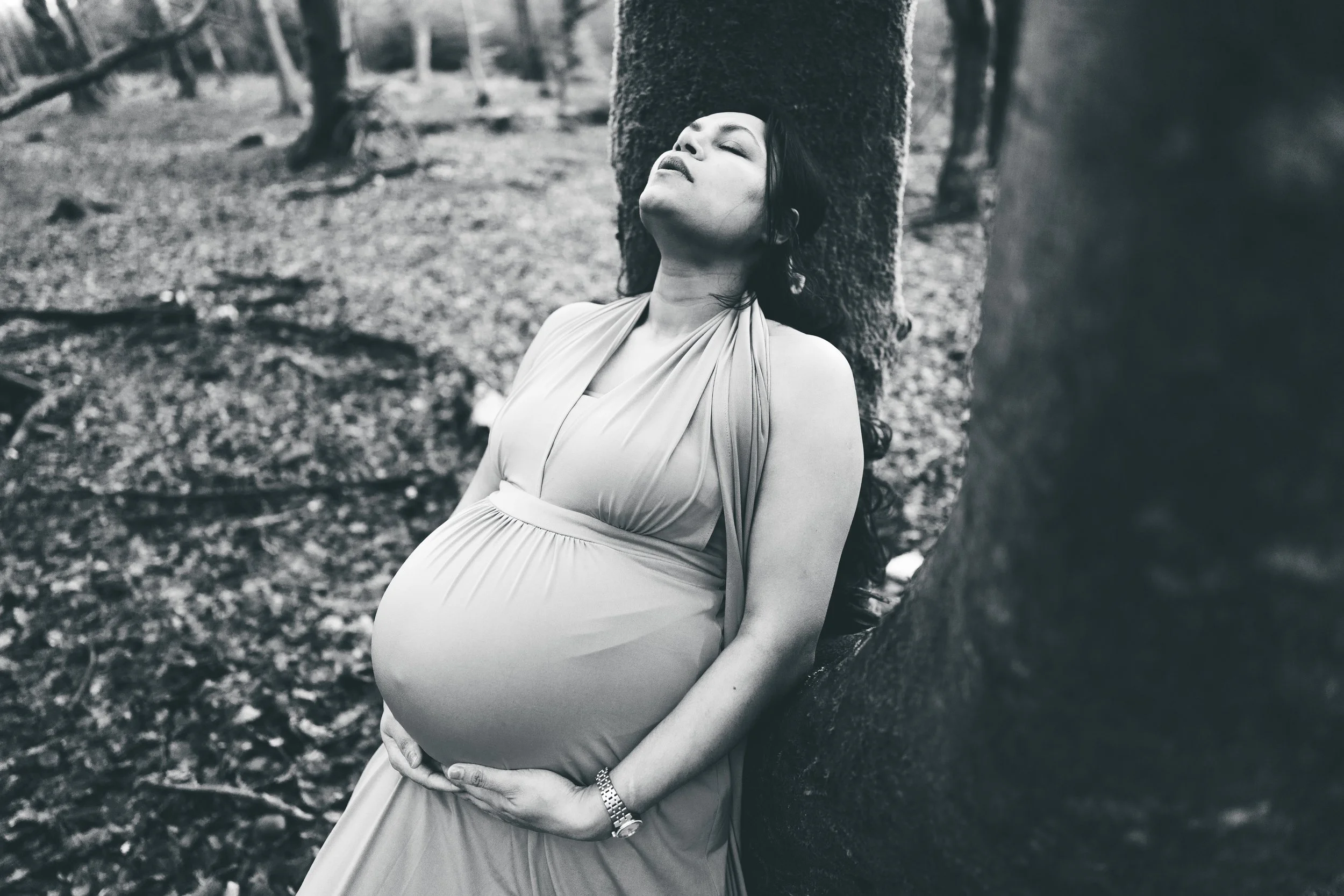 Black and white photo of a pregnant woman leaning against a tree in a wooded area, with her eyes closed and head tilted back.