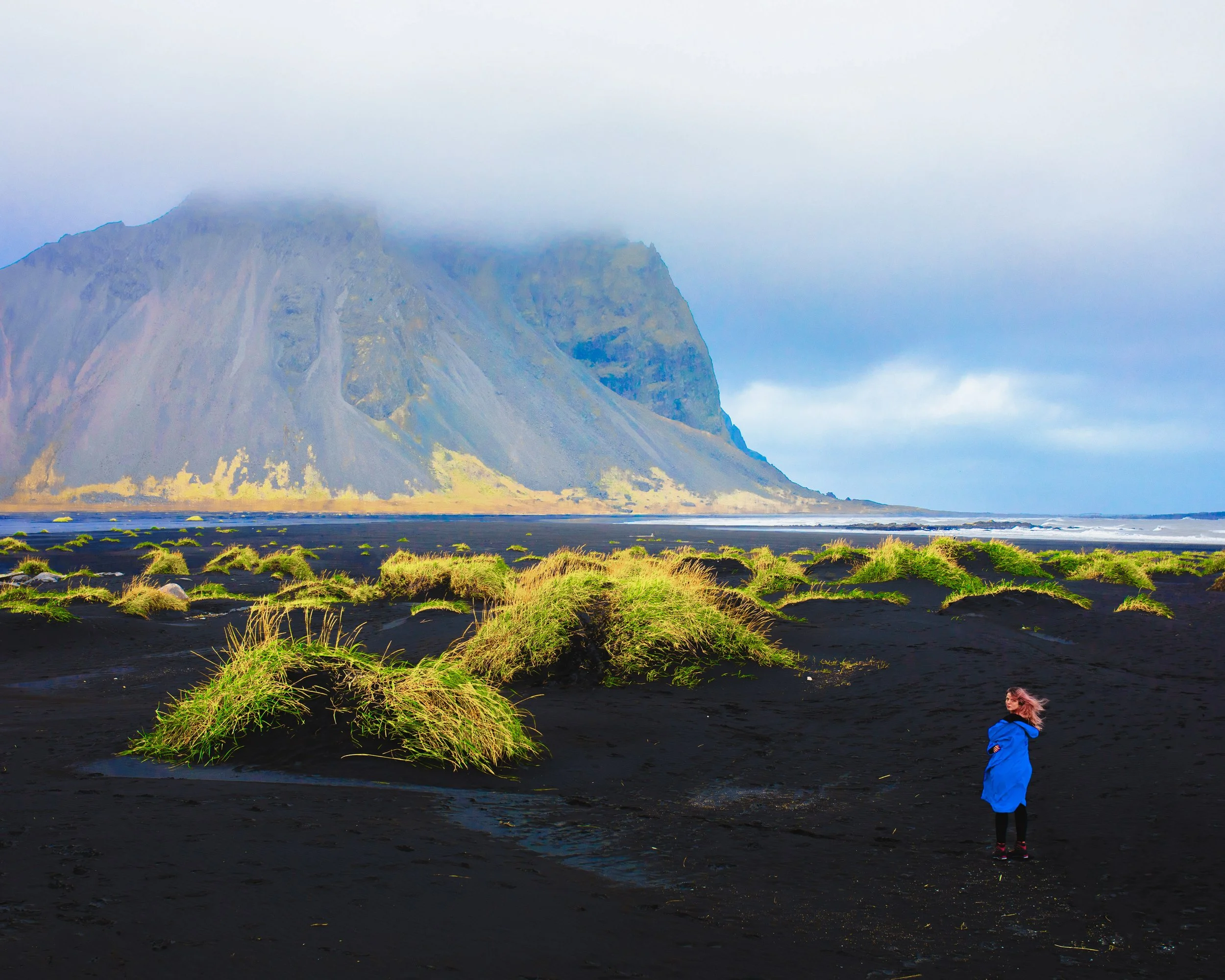 Woman standing on black sand dunes with grassy mounds and large mountain behind