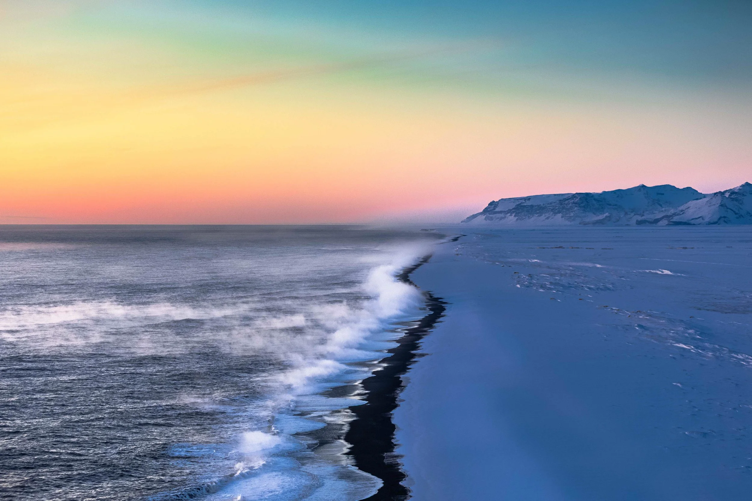 Pink hour colours over an Icelandic coastline with blue snow and waves breaking along a black sand beach