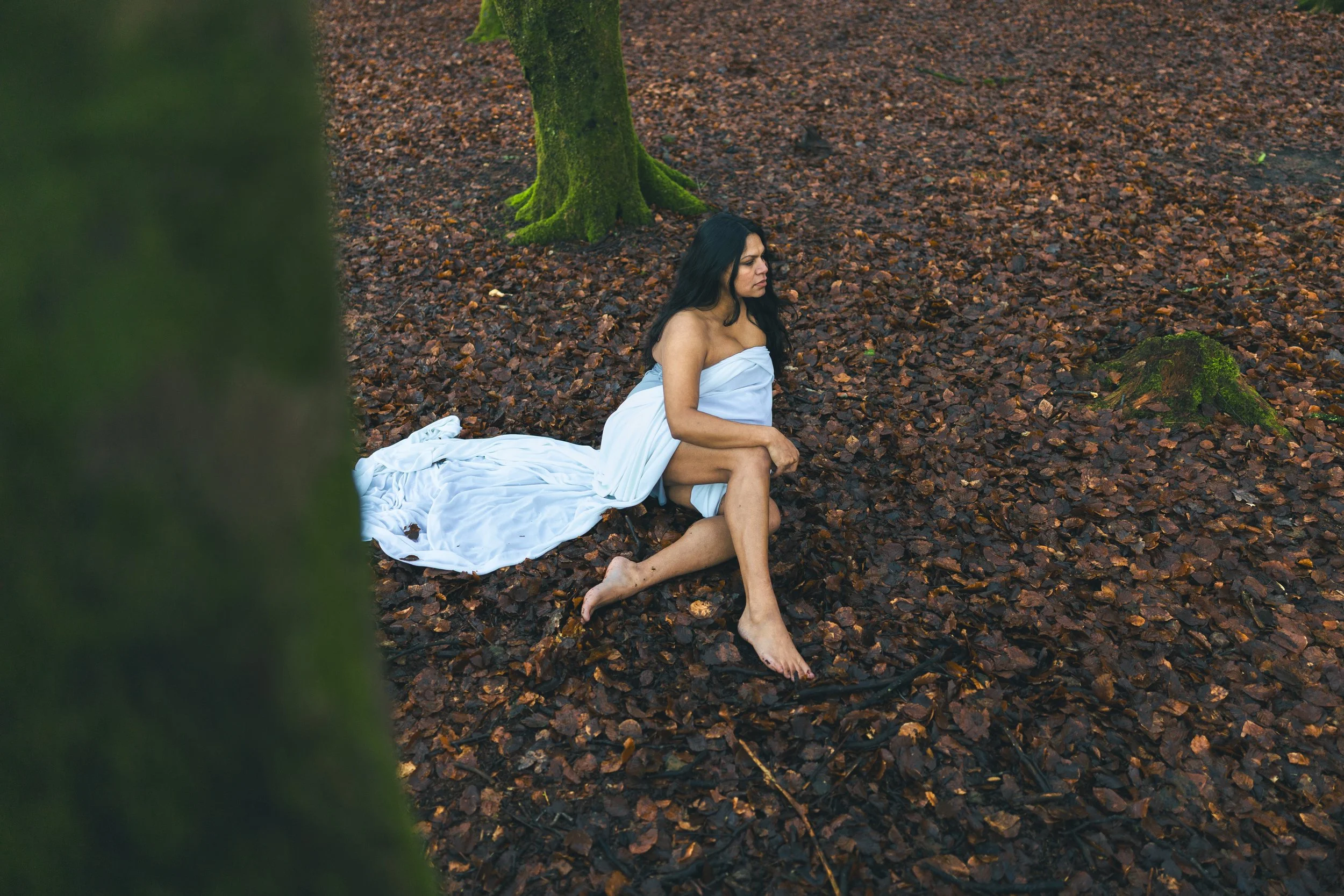 A woman with long dark hair wearing a white towel sits on wet brown leaves in a forest, holding her knees with a contemplative expression.