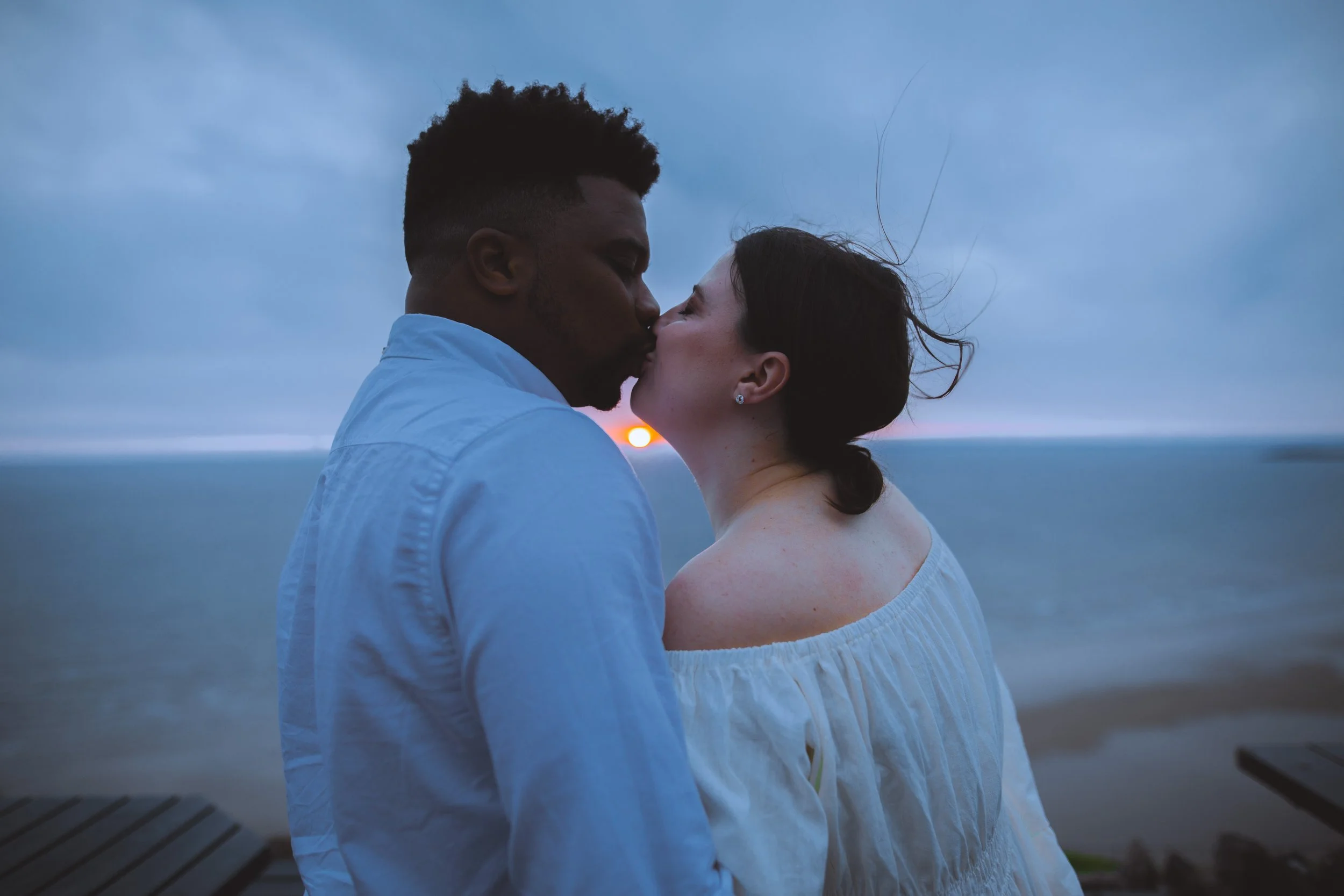 Couple kissing outdoors at sunset with the ocean in the background.