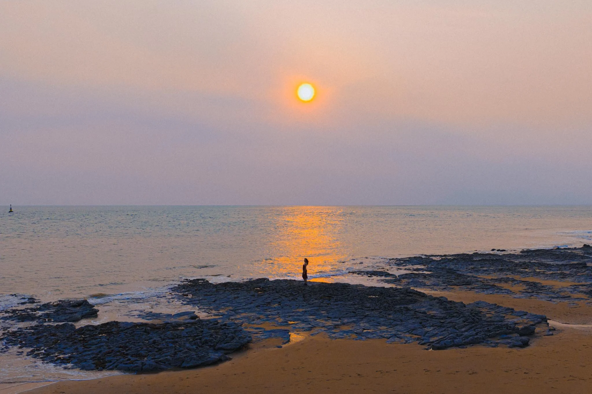 Woman standing on rocky shoreline at sunset with reflection of sun on calm sea