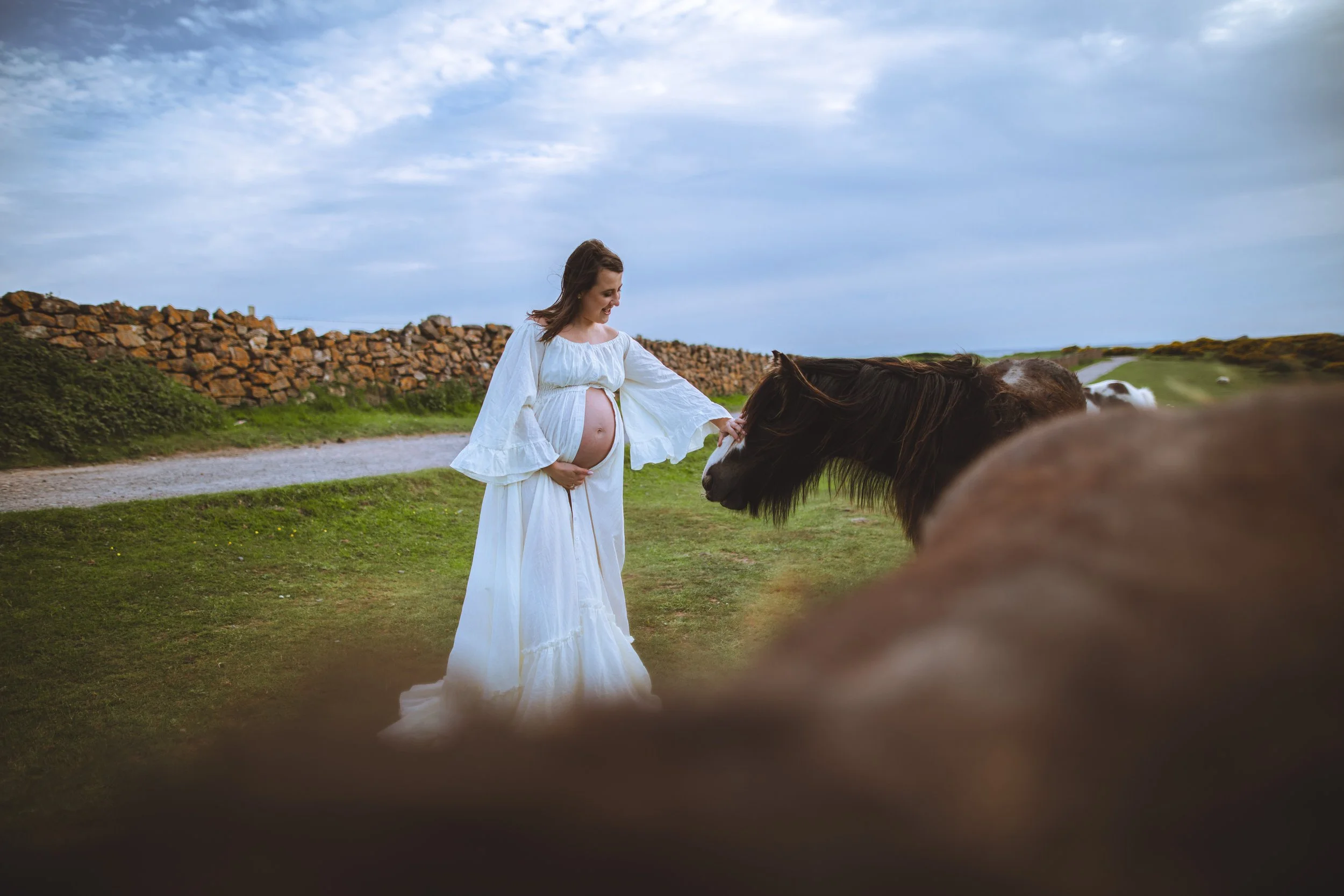 A pregnant woman in a white dress gently touching the nose of a horse outdoors on a grassy area with a stone wall and cloudy sky in the background.