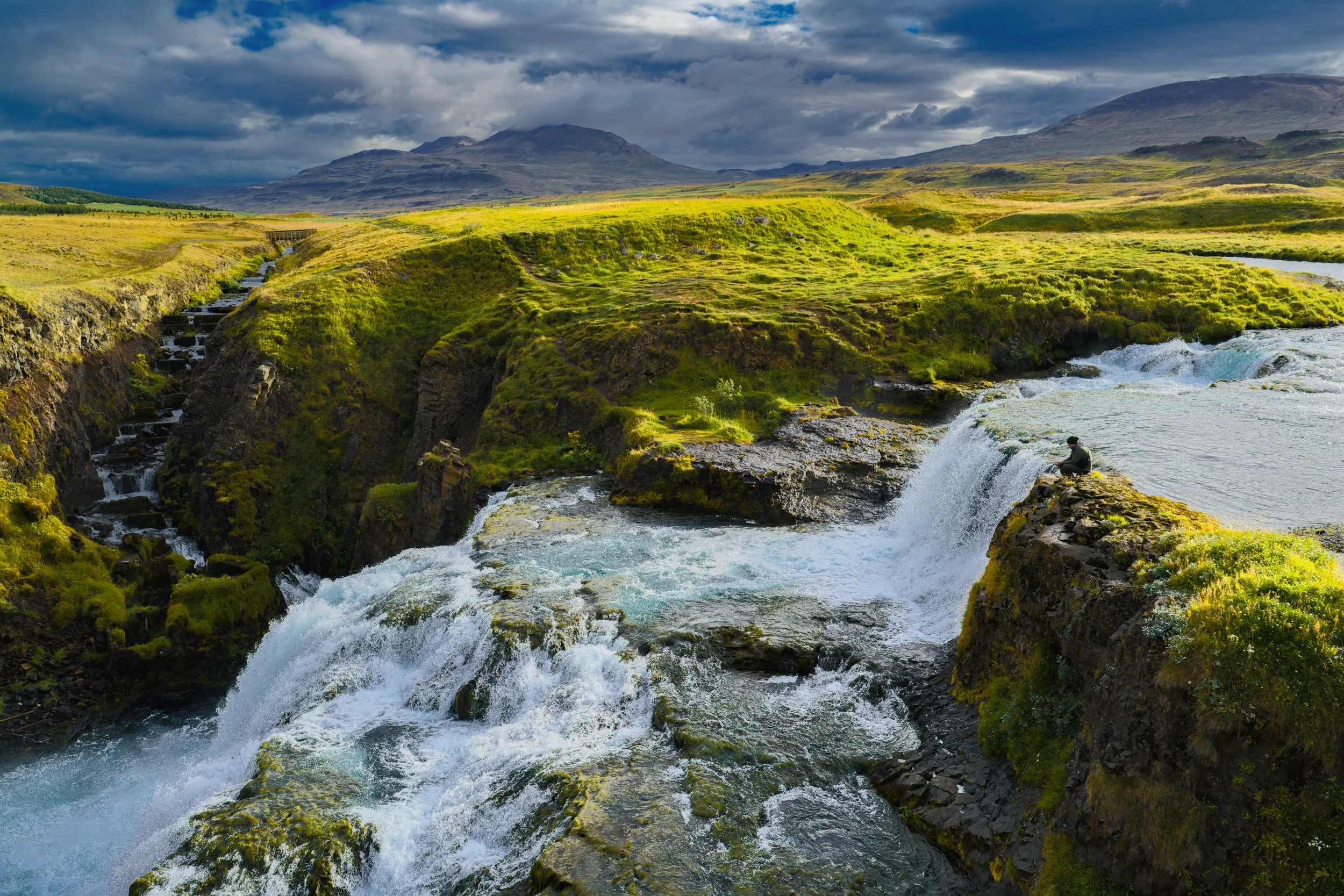 Panoramic Icelandic landscape with a cascading waterfall, moss-covered cliffs, green meadow, rugged mountains, and a person sitting beside the falls.