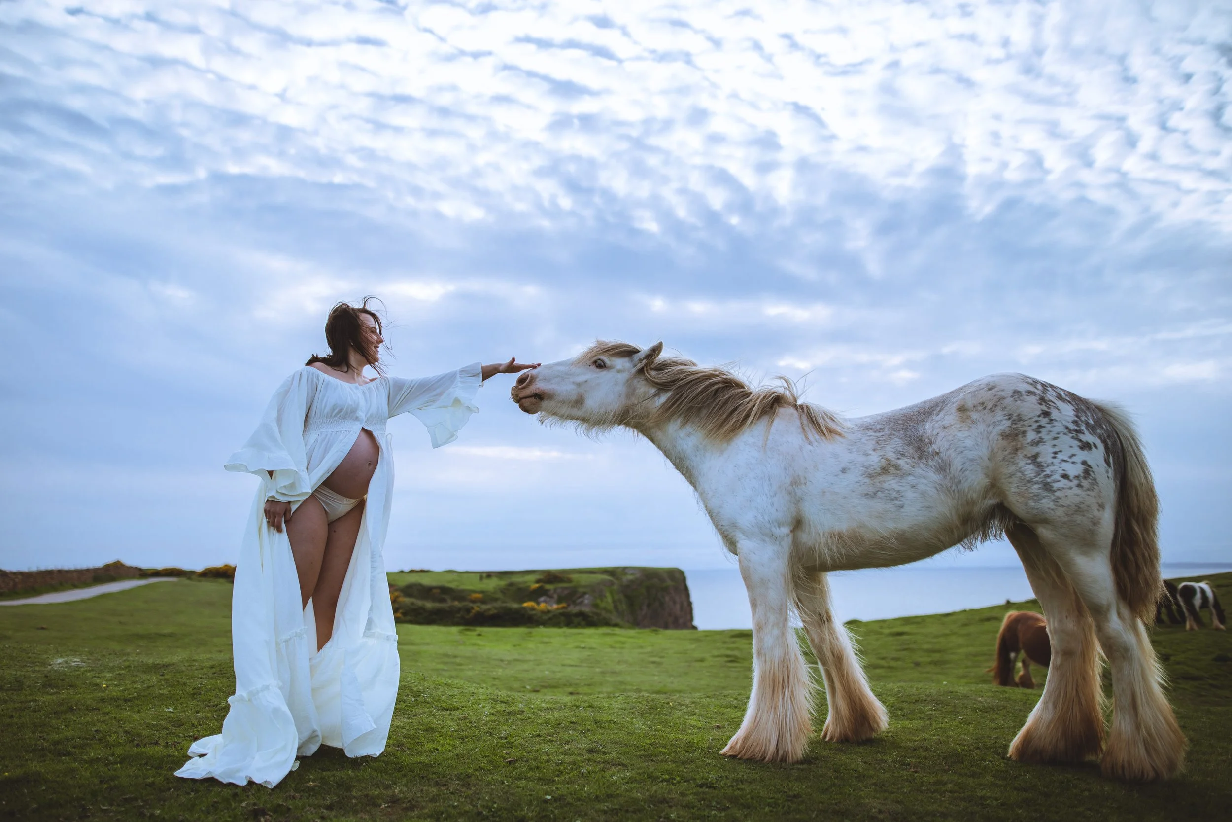 A woman with long hair and a flowing white dress touching a white horse with brown spots on a grassy landscape under a cloudy sky.