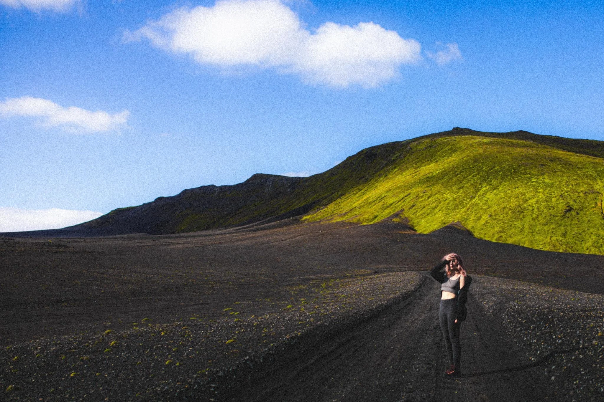 Woman standing on black sand path with green hills rising behind under blue sky