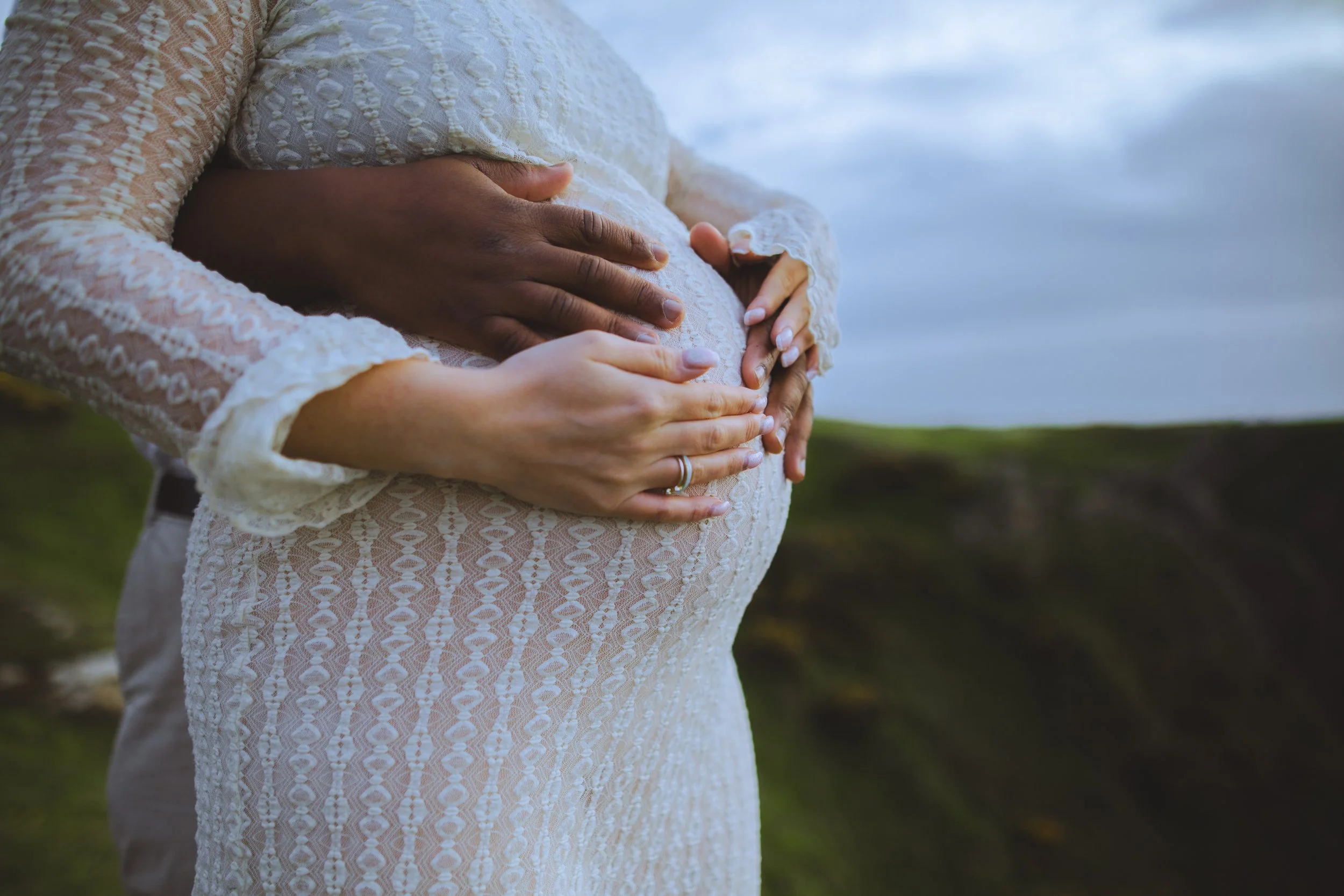 Close-up of a pregnant woman in a white lace dress with hands holding her baby bump, with another pair of hands on top, outdoors with a cloudy sky and green landscape in the background.