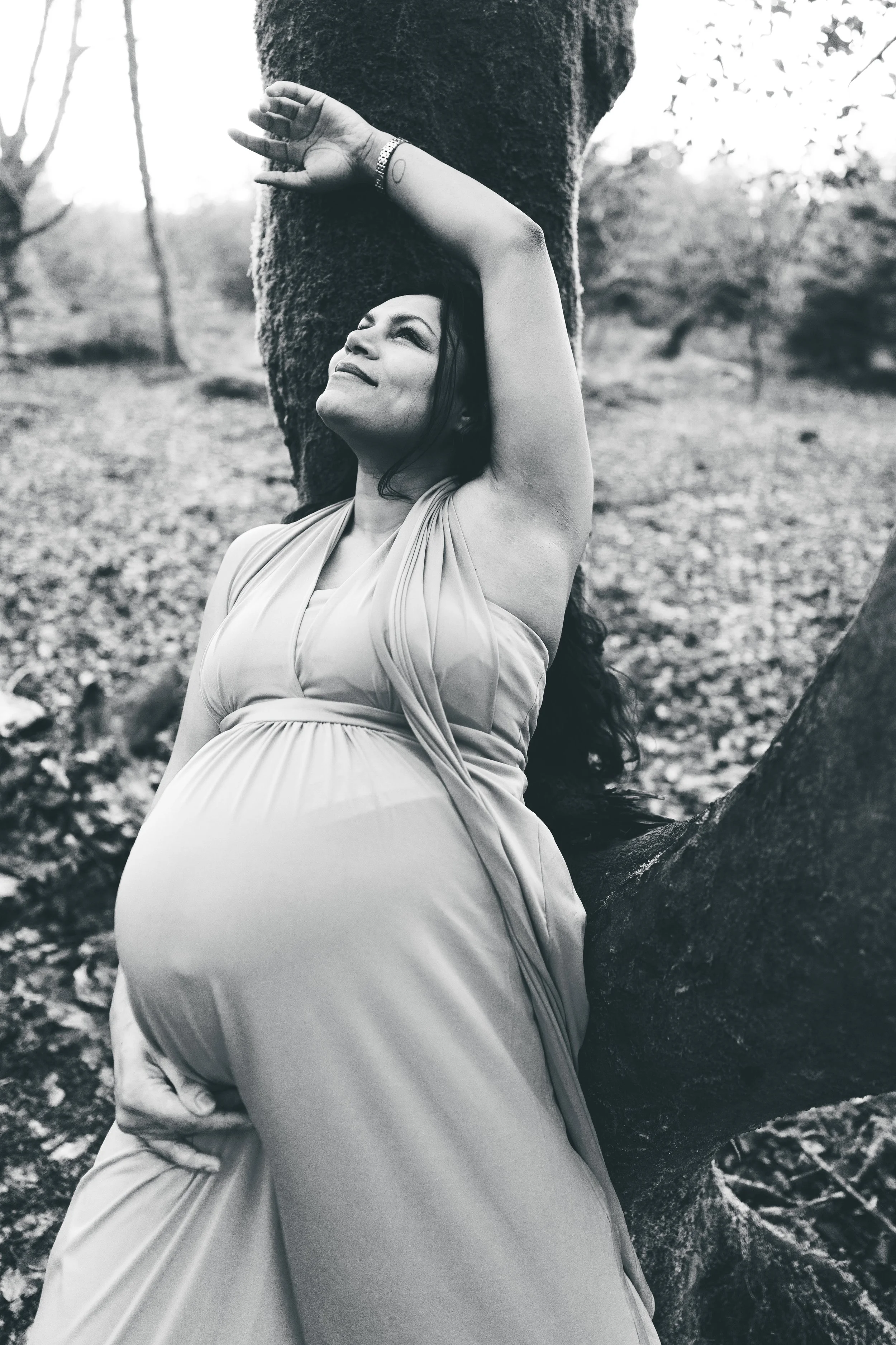 A woman in a flowing dress leaning against a tree in a forested area, with a calming expression on her face and one arm raised above her head.