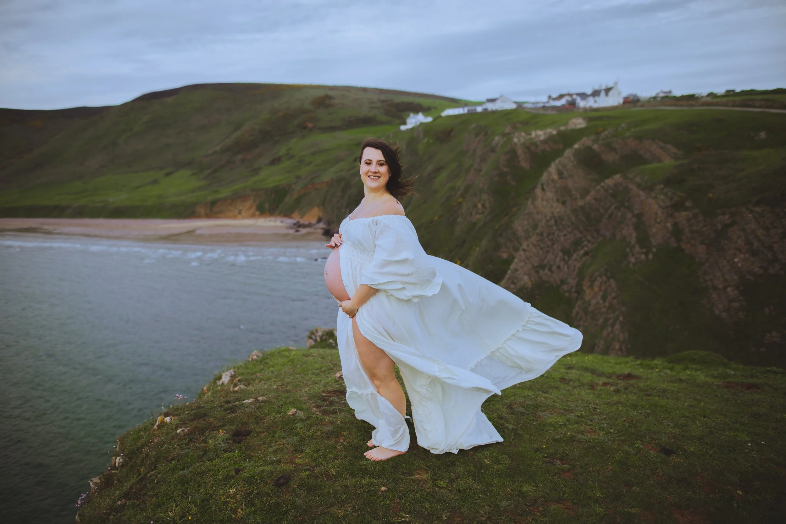 Pregnant woman in white dress standing on grassy cliff overlooking ocean and cliffs in background