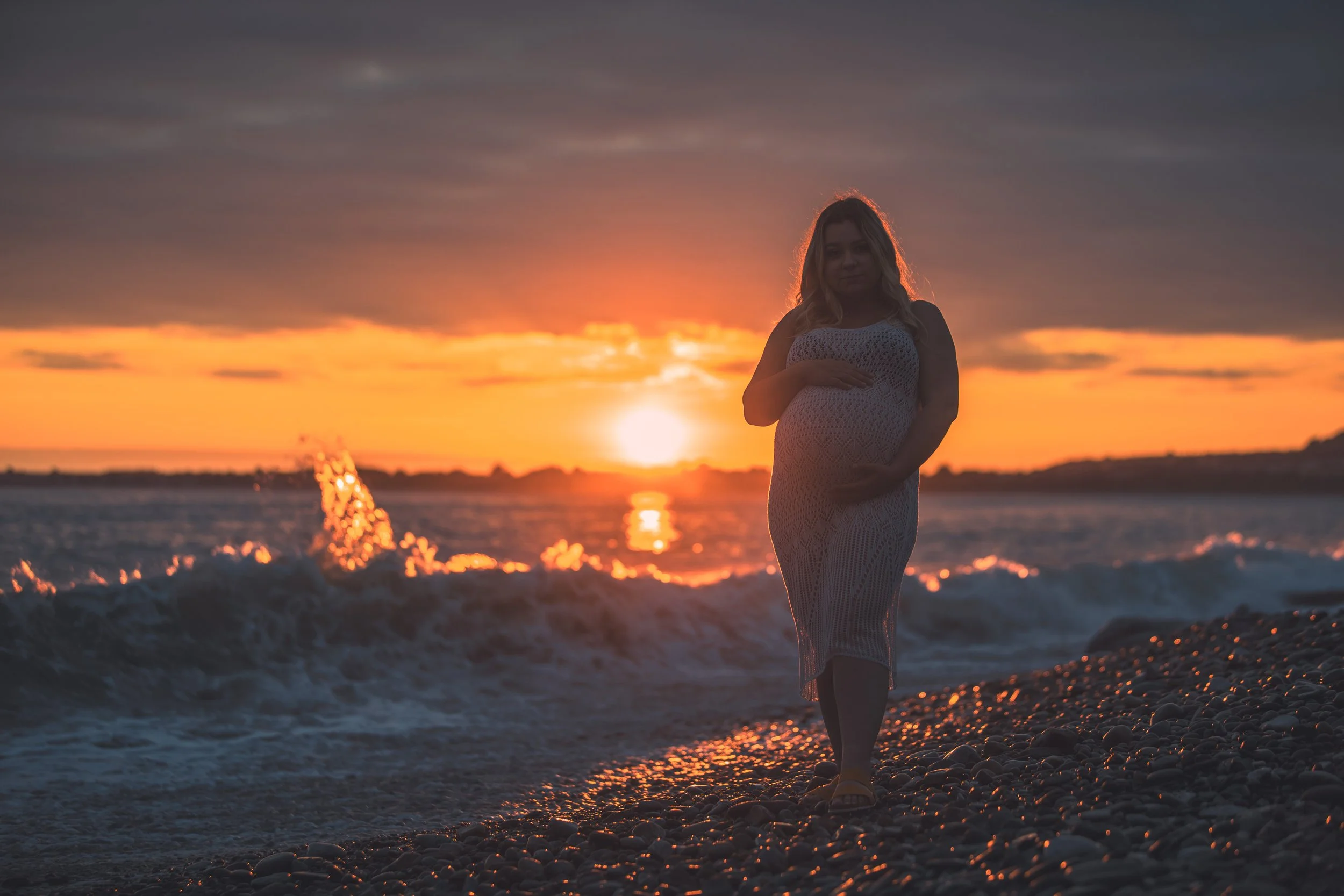 A woman walking on a pebbled beach during sunset, with her hand resting on her pregnant belly, wearing a white crochet dress.