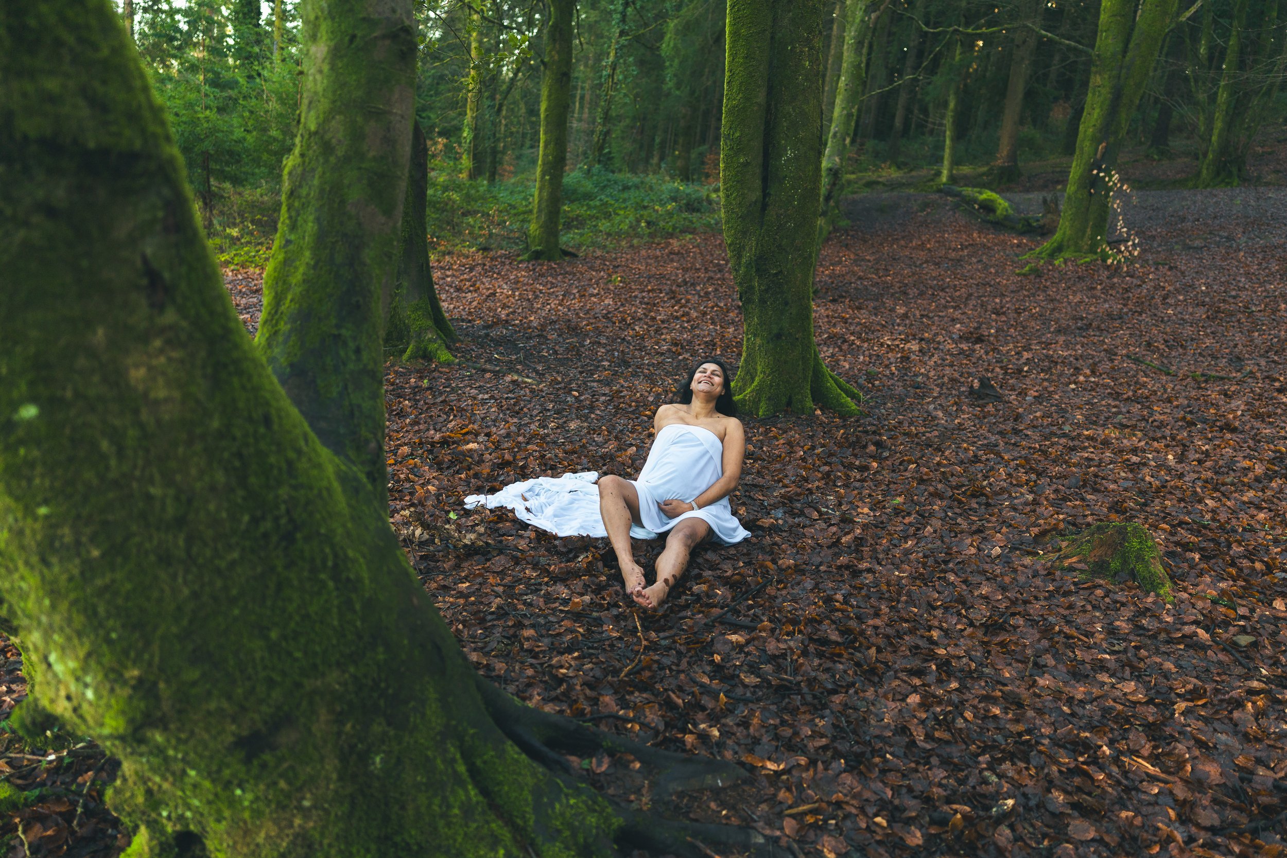 A woman wearing a white dress sitting on the forest floor, smiling and looking up, surrounded by tall moss-covered trees in a lush green forest.