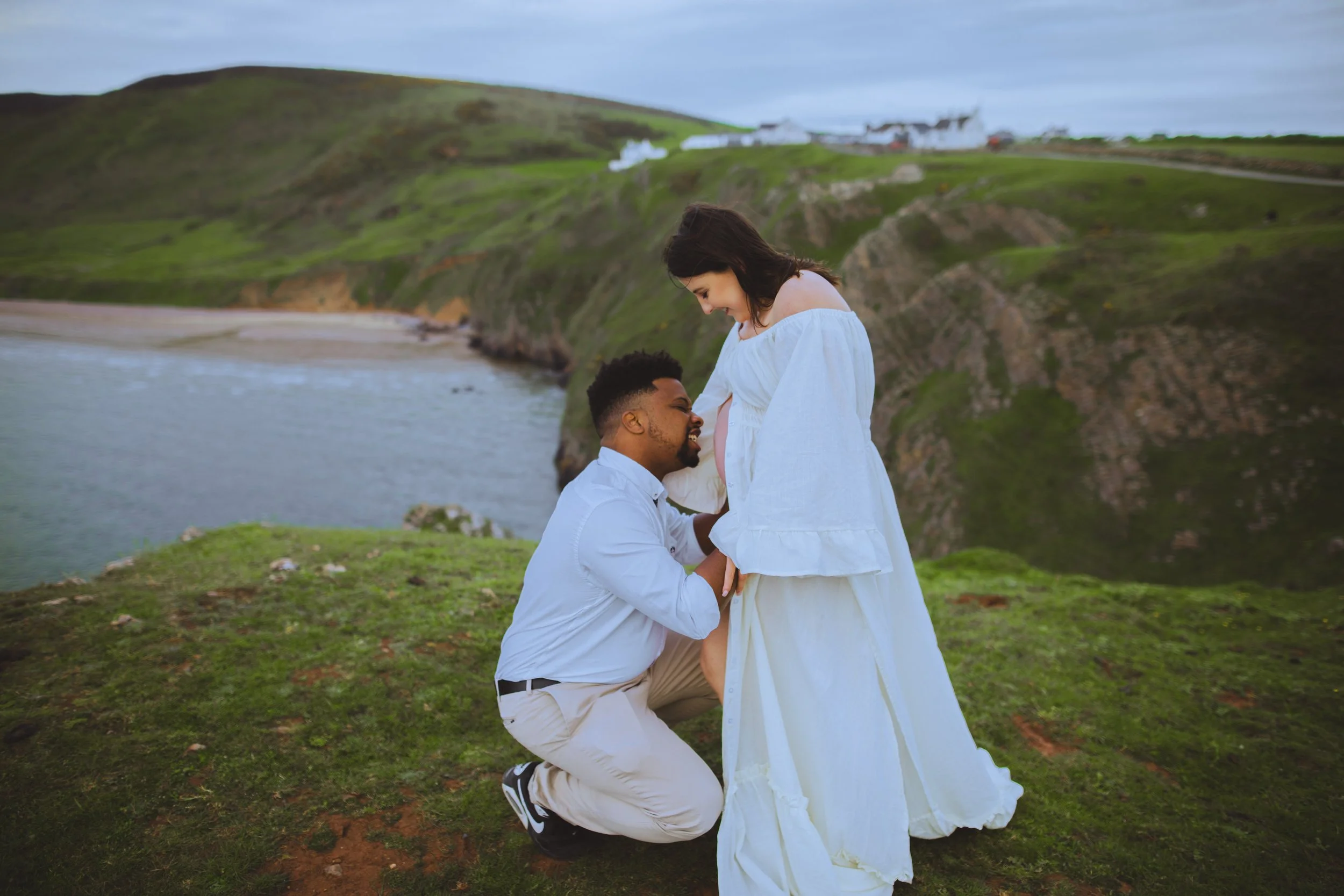 A man kneeling on grass, holding and smiling at a pregnant woman while she looks down at him, near a coastline with cliffs and water in the background.